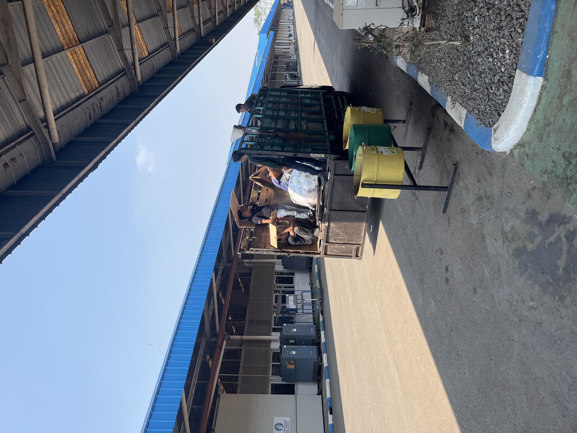 The image depicts a scene outside a building with a blue roof, possibly a warehouse or factory, in Pelang, Indonesia. A truck with a green cargo area is parked, and several men are engaged in loading or unloading items from it. In the foreground, there are yellow and green trash bins. The ground is asphalt, with a shadow cast by an overhead structure. The environment appears to be an industrial or commercial area, characterized by the large building and the presence of a delivery truck. The sky is clear and blue.