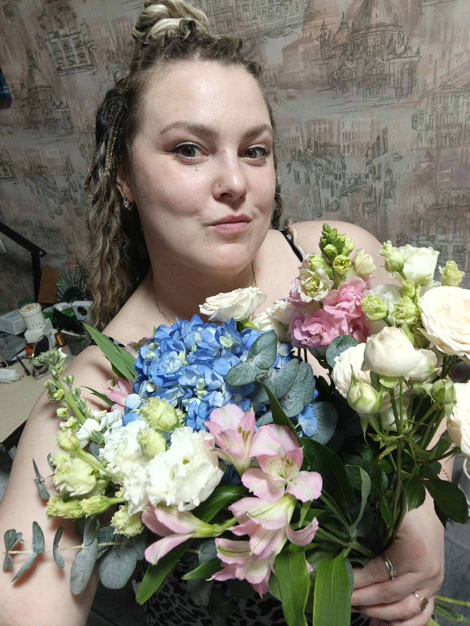A medium shot captures a woman holding a bouquet of flowers, likely indoors. The woman has fair skin, brown eyes, and curly hair with some braided strands. Her hair is styled in a partial updo. She is wearing a printed top. A small nose ring adorns her nose. The bouquet is diverse, featuring blue hydrangeas, pink alstroemeria, white roses, and green stock. The flowers are tightly packed together, obscuring the woman's torso. In the background, the wallpaper has a detailed architectural scene, possibly depicting European cityscapes. The left side of the frame reveals a cluttered surface with various objects. The exact location, Марковский, Russia, is not evident from visual details.
