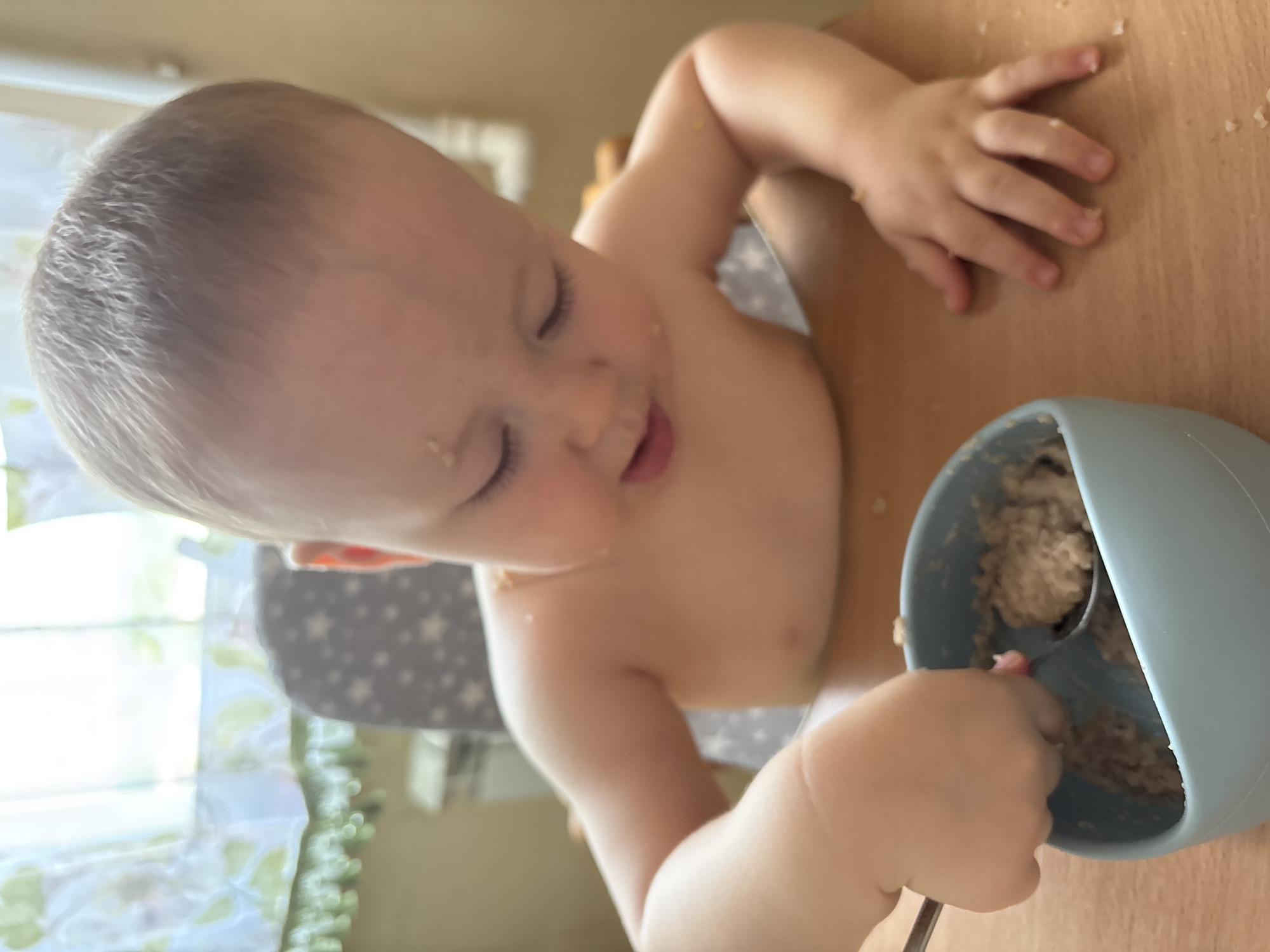 A shirtless baby is seated in a high chair at a wooden table, actively eating from a light blue bowl with a spoon. Oatmeal or a similar food is present in the bowl and scattered on the baby's face, chest, and the table surface. The background shows a window with sheer curtains and a glimpse of what appears to be a kitchen or dining area, suggesting a domestic setting. The lighting suggests it is daytime. There is no information to determine the location as Naberezhnye Chelny, Russia.