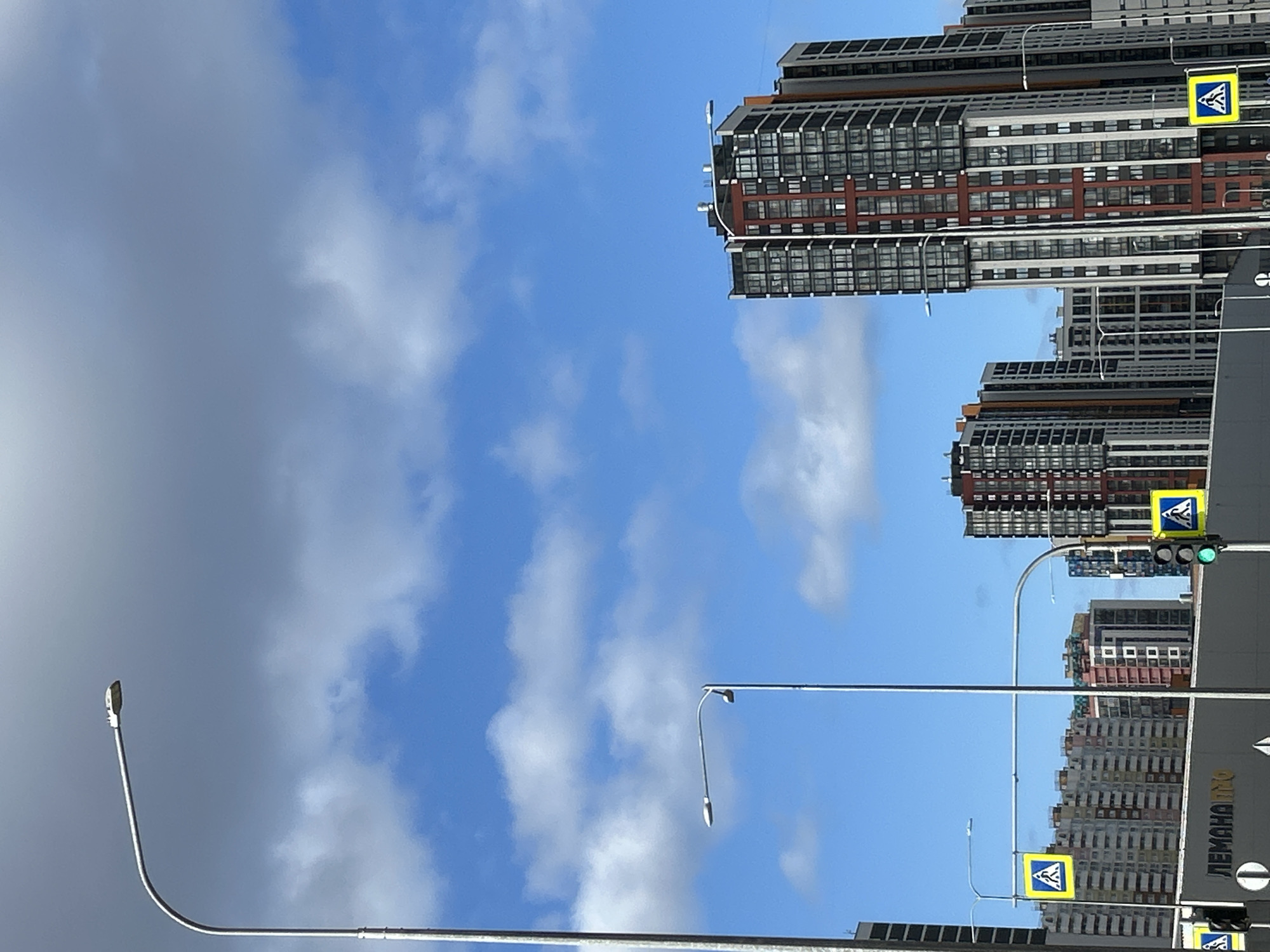 The image shows a view of a city, likely Saint Petersburg, Russia, based on the architectural style and the Cyrillic lettering on signage. Tall, modern apartment buildings with repetitive window patterns dominate the skyline. The sky is partly cloudy with patches of blue. Streetlights line the road, and pedestrian crossing signs are visible. Traffic lights are present, with one showing a green light. A significant portion of the image is occupied by the sky and clouds.