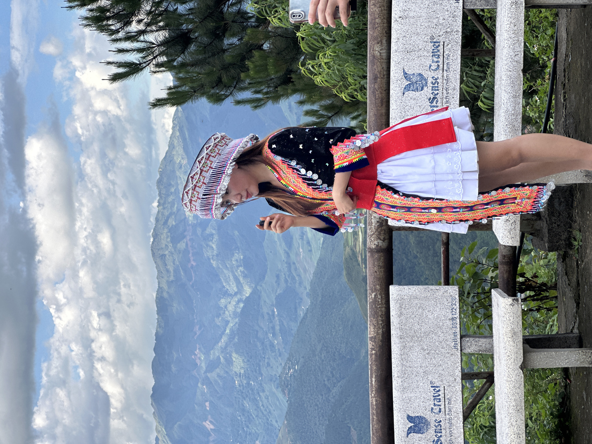 A woman in traditional attire stands outdoors at a scenic viewpoint in Tu Le, Vietnam, with a mountainous landscape in the background. The woman, positioned slightly right of center, faces left with her head tilted slightly up. She wears an elaborate headpiece featuring intricate white, red, and black patterns with numerous dangling silver-colored discs. Her long-sleeved black top is adorned with colorful embroidery in red, yellow, and blue, and also features silver-colored disc embellishments. She has a bright red sash around her waist and a white, pleated skirt. A long, colorful embroidered panel, matching the style of her top, hangs down her left side. In her right hand (viewer's left), she holds a small, dark, cylindrical object. To her right (viewer's right), a hand holding a silver-colored smartphone with a multi-lens camera array is visible, pointing towards her, indicating she is being photographed.
In the immediate foreground, a rustic metal railing separates the viewer from the scene. Behind the railing are two speckled gray granite benches. The bench on the left displays the logo and text "Sense Travel®" along with partially visible Vietnamese text "lich voi niem dam me" and a hotline number "0916 172 338". The bench on the right also shows the "Sense Travel®" branding.
The background consists of extensive, verdant mountains covered in dense foliage, with visible terraced fields and scattered structures on the slopes. The sky above is partly cloudy, featuring large white and gray cumulus clouds with patches of blue sky. A tall pine tree with long needles is partially visible on the far right. Green shrubs and plants grow behind the benches and railing.