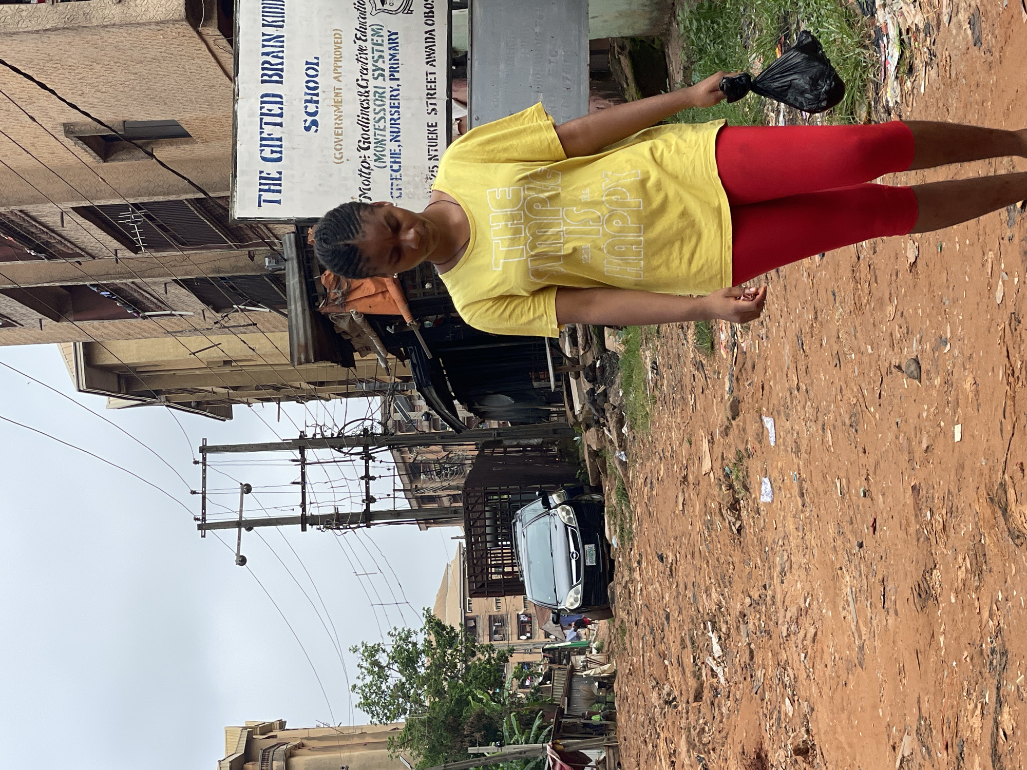 The image shows a young woman standing on a red dirt road in Nkpor, Nigeria. She is wearing a yellow t-shirt with the phrase "The Simple is Happy" written on it and red leggings, and she is holding a black plastic bag. Behind her is a street scene with buildings, power lines, and a parked black vehicle with license plate "AGU-GZSAE." There is a sign for "The Gifted Brain Kid School," which is "Government Approved" and has a motto of "Godliness & Creative Education (Montessori System)" and offers creche, nursery, and primary education at "5 Ntueke Street, Awada Obosi". The road appears to be littered with trash.