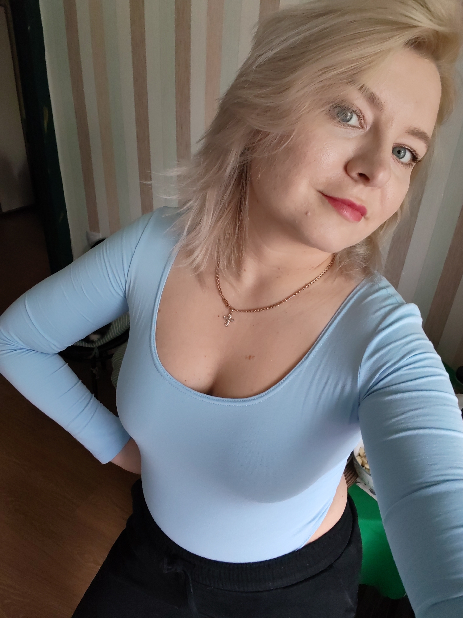 An indoor, high-angle self-portrait features a young adult woman with shoulder-length blonde hair and blue eyes, looking directly into the camera with a slight, closed-lip smile. She has fair skin, a small mole below the right corner of her mouth, and is wearing red lipstick. Her attire consists of a form-fitting, light blue, long-sleeved top with a low scoop neckline, revealing a gold chain necklace with a small cross pendant. She is also wearing dark (likely black) casual pants with an elasticated waistband, visible at the bottom of the frame. Her left hand is placed on her hip.
The background consists of a wall featuring vertical stripes in alternating shades of light cream, beige, and light brown. To the far left, a dark green vertical element, possibly a curtain or doorframe, and a dimly lit doorway are visible. The bottom left foreground shows a light brown wooden floor. A blurred object, potentially a bowl containing light-colored items, is partially visible on the far right side of the frame. The lighting is soft and even, consistent with an indoor environment.
Based solely on the visual information provided in the image, there are no discernible features, architectural elements, or contextual cues that indicate the specific location is the city of Izhevsk, Russia.