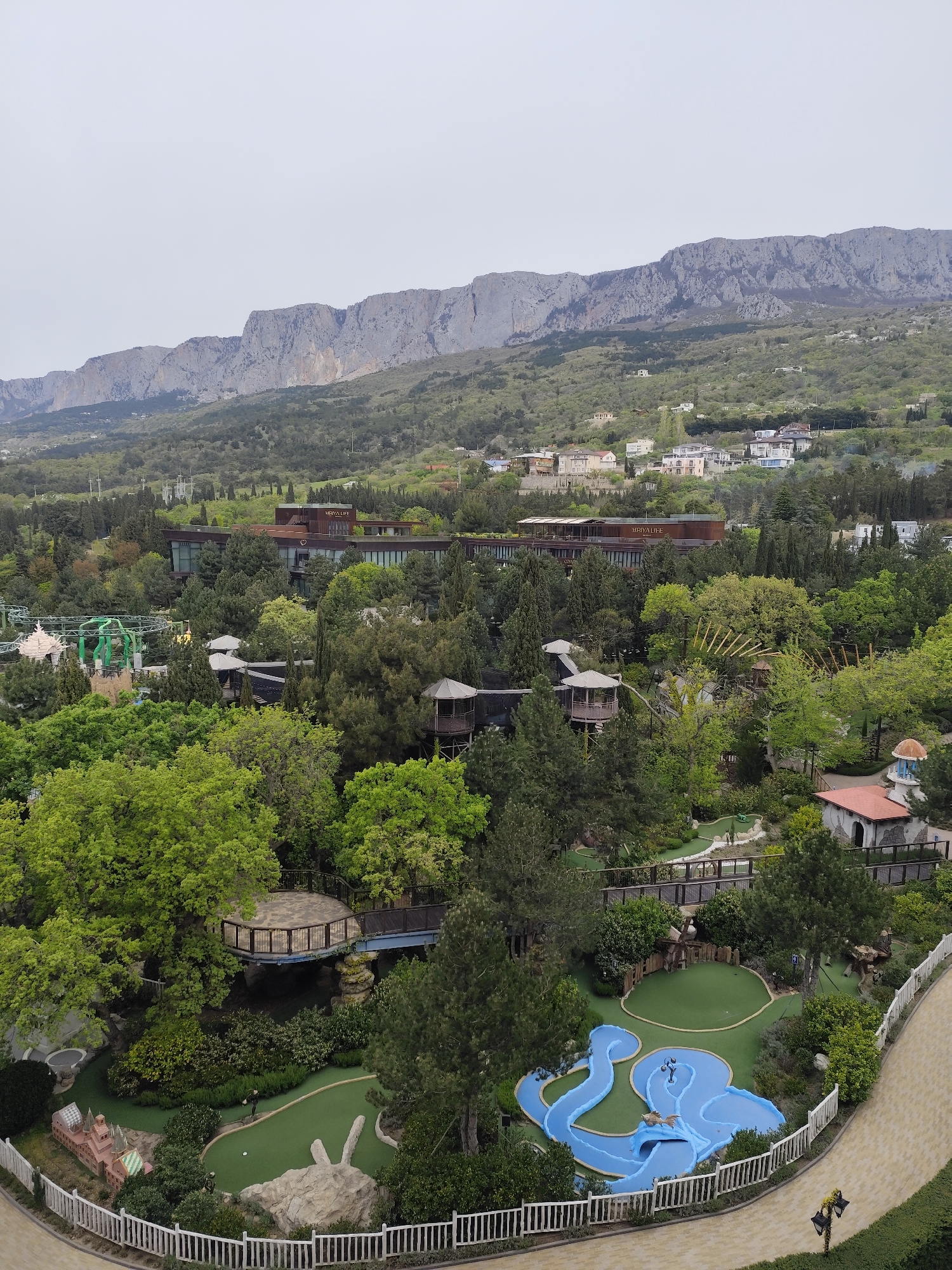 This image presents a panoramic view of a lush, mountainous resort area, likely within the Симеизский поселковый совет, Ukraine. The scene is dominated by dense green foliage, with a towering, rugged mountain range forming a dramatic backdrop under an overcast sky.
In the immediate foreground, a paved walkway with a white picket fence curves through a recreational area. Several mini-golf courses are visible, featuring manicured green turf, landscaping, and decorative elements. One prominent course includes a winding, multi-level blue water play feature. Small figures of people can be seen on the mini-golf courses, engaged in play.
The midground is a sprawling park-like setting, heavily wooded with a mix of deciduous and coniferous trees. Nestled among the trees are various wooden structures, including elevated gazebos or treehouses connected by bridges. A green roller coaster track is visible winding through the greenery, suggesting an amusement park component. A large, modern building with extensive glass windows and a reddish-brown facade stands out amidst the trees, with the text "MRIYA LIFE" clearly visible on its side.
Further up the hillside, beyond the main resort complex, residential buildings with light-colored roofs are scattered among the trees. The background is dominated by the majestic, grey rock formations of the mountain range, which stretches across the horizon. The overall impression is one of a vast, well-maintained leisure and recreation destination set against a stunning natural landscape, captured during daytime under a cloudy sky.