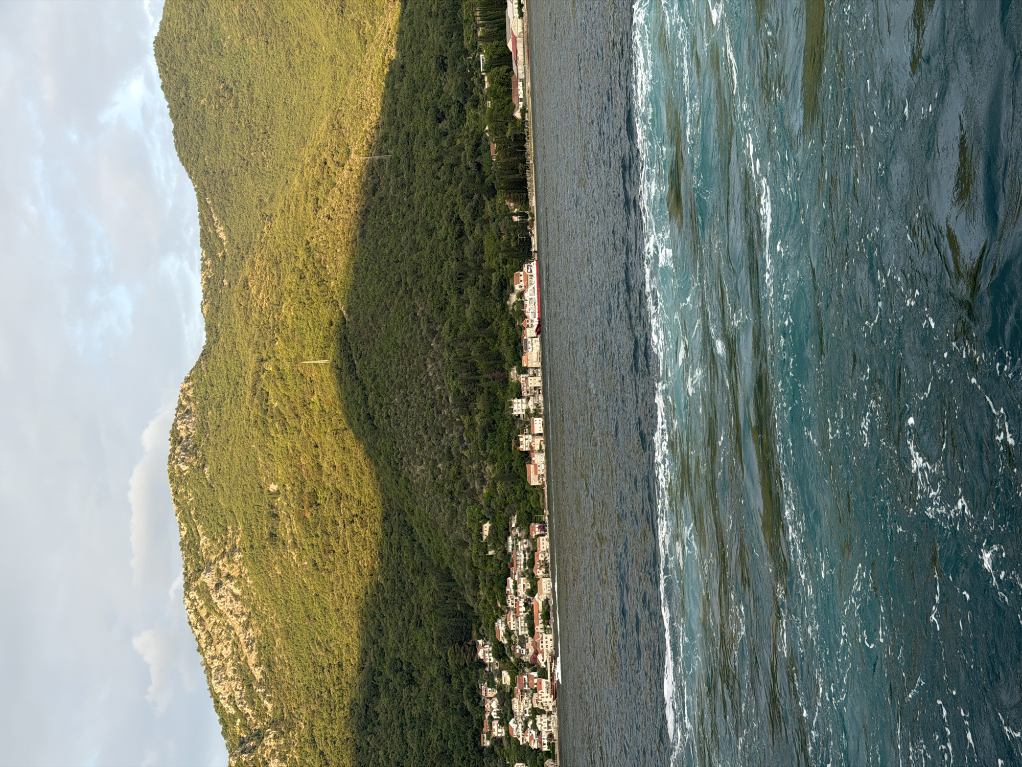 This image depicts a body of water in front of a coastal town nestled at the foot of a lush, green mountain. The town consists of buildings with white walls and red roofs, clustered together along the shoreline. The mountain behind the town is densely covered with trees, with rocky outcrops visible in some areas. The sky above is partly cloudy, with sunlight casting a warm glow on the mountain and the town. The water in the foreground is choppy, with white foam indicating movement, possibly from a boat or a strong current. The scene appears to be a peaceful, scenic view of a coastal settlement in Đurići, Montenegro.