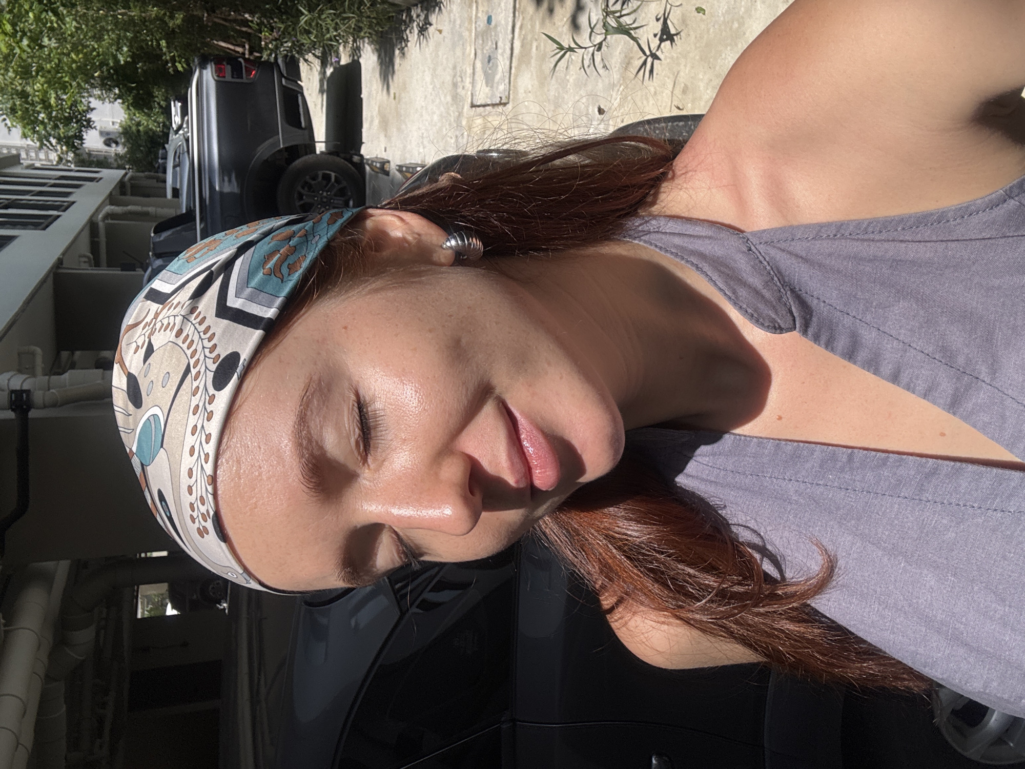 A woman is seen in a close-up shot, likely taken in a parking area. She is wearing a patterned headscarf and a gray top. A dark-colored SUV is parked behind her. The setting suggests an urban environment in daylight, possibly within the city of Choeng Thale, Thailand. The scene has natural sunlight with a slight glare.