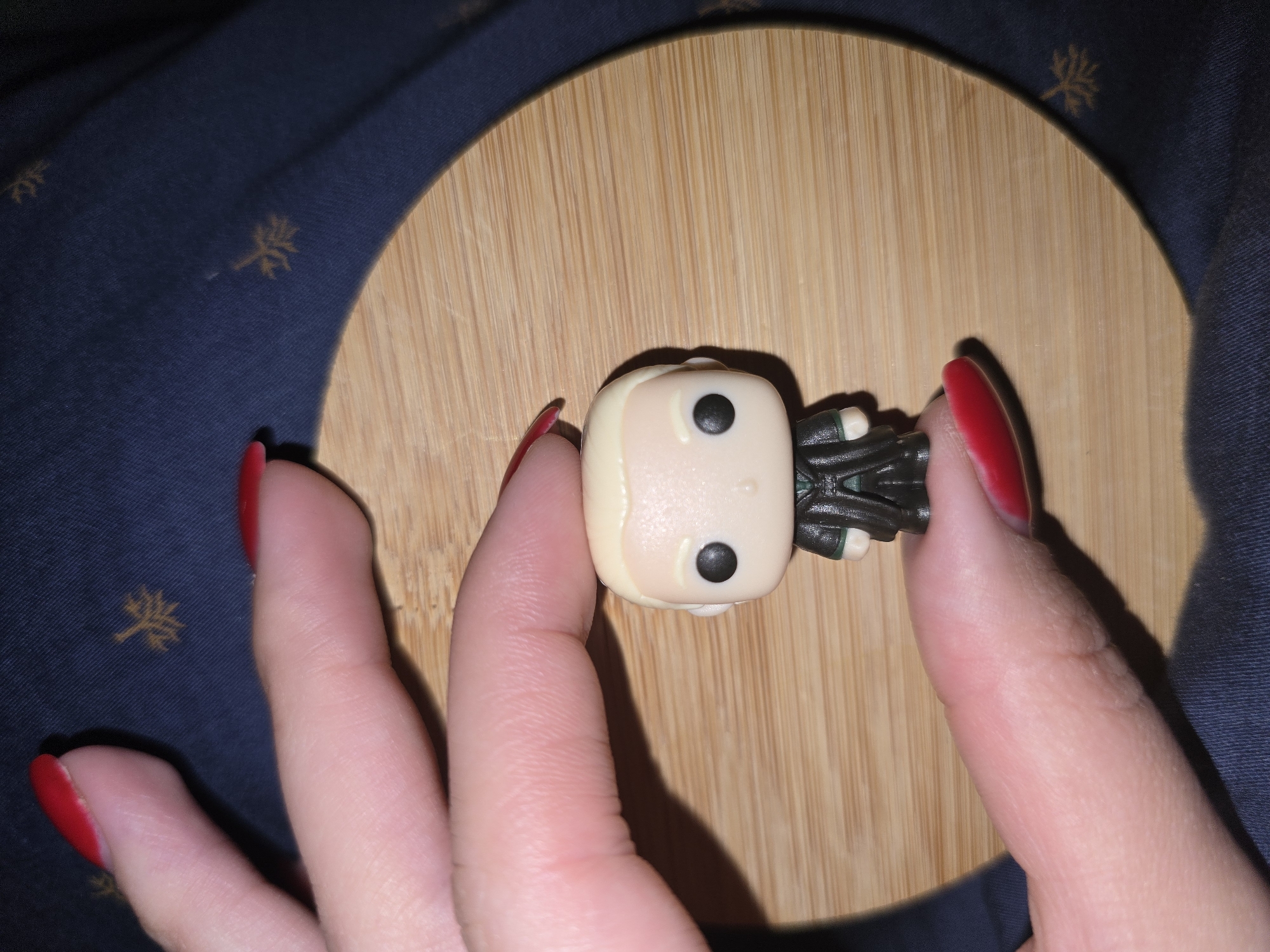 A close-up, overhead shot shows a person's hand with bright red fingernails holding a small, blonde-haired, Funko Pop-style figurine wearing a dark green robe. The figurine is positioned on a round, light brown wooden surface. The background is a dark blue fabric with a subtle, gold-colored, snowflake-like pattern. The scene is static, with no apparent action or interaction occurring. The image is lit to highlight the figurine and the hand. No specific location in Petropavlivska Borshchahivka, Ukraine can be determined from the visual information provided.