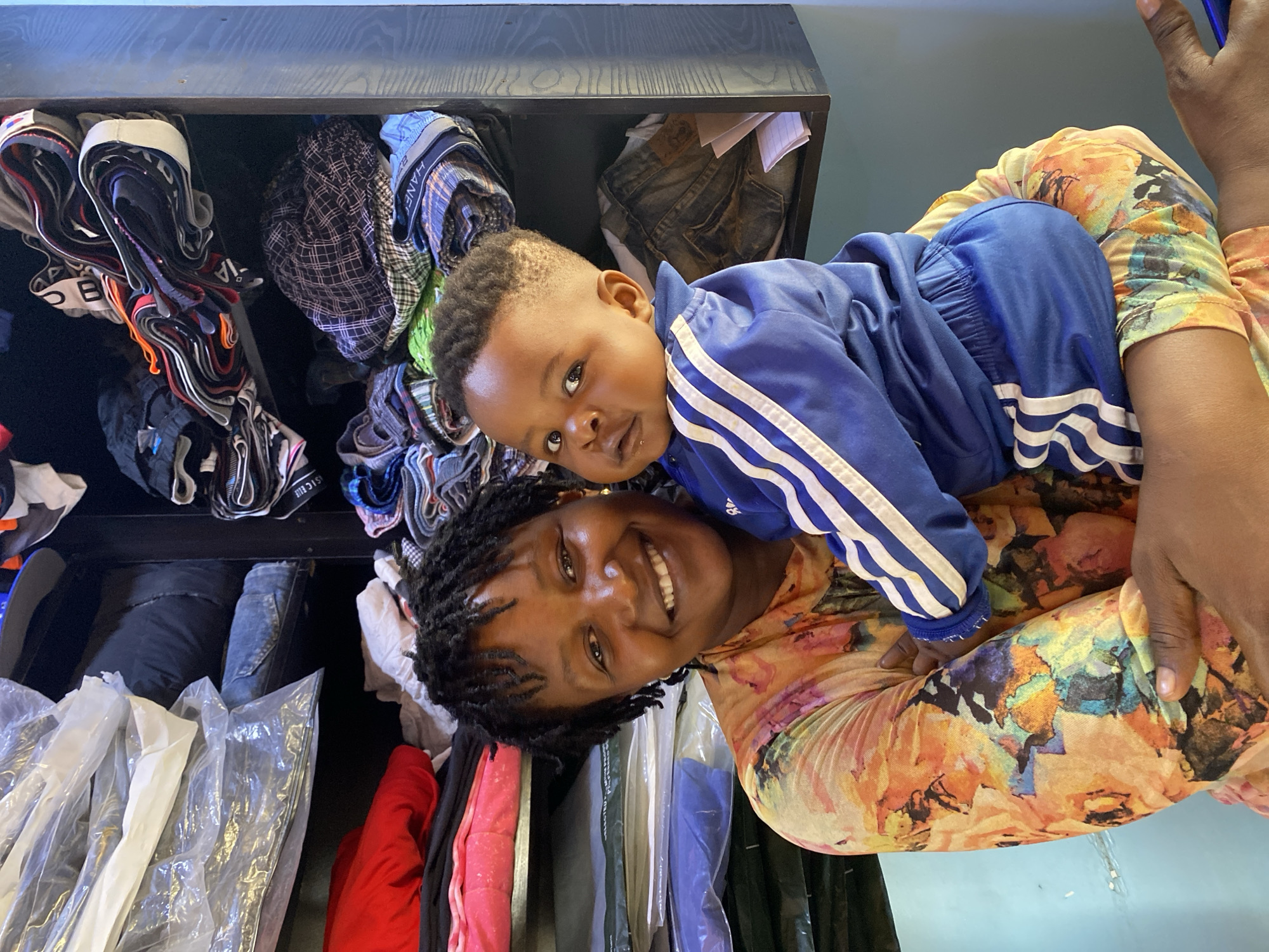 A woman with dark skin and dark hair smiles while holding a baby wearing a blue tracksuit in an indoor setting. Behind them is a closet with clothes. The specific location is Bukuru, Nigeria, given the context.