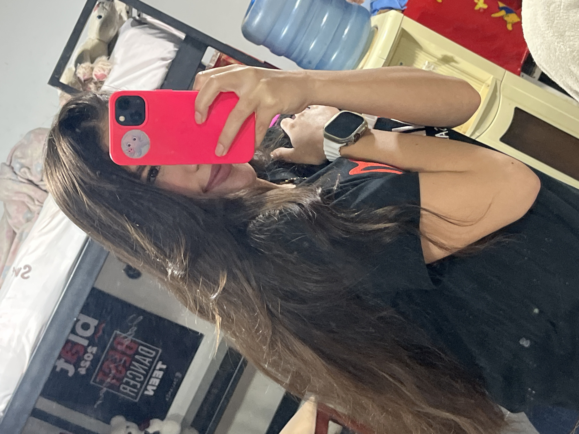 The media file shows a person taking a selfie in a bedroom. The person, likely female due to the long hair and pink phone case, is lying down and holding up a pink smartphone to capture their reflection in a mirror. The person is wearing a black t-shirt with an orange design on it and a white watch.
The bedroom appears to be somewhat cluttered. In the background, a large blue water jug is visible, along with a cream-colored dresser or cabinet. A red blanket with yellow motifs is draped over the furniture. A stuffed animal, possibly a bear, is partially visible on a bed with white bedding.
The lighting suggests it is daytime, but the specific time is not clear. The environment seems to be indoors, a private living space. The person's expression is not fully visible, but the act of taking a selfie implies a casual or playful mood.
Text visible in the reflection on a poster or book reads "TEEN BEST DANCER" and "ASOS PLAY". The poster is positioned vertically and appears to be dark blue with white and red lettering.
The overall impression is of a candid moment captured in a personal space, with the focus on the person and their immediate surroundings.