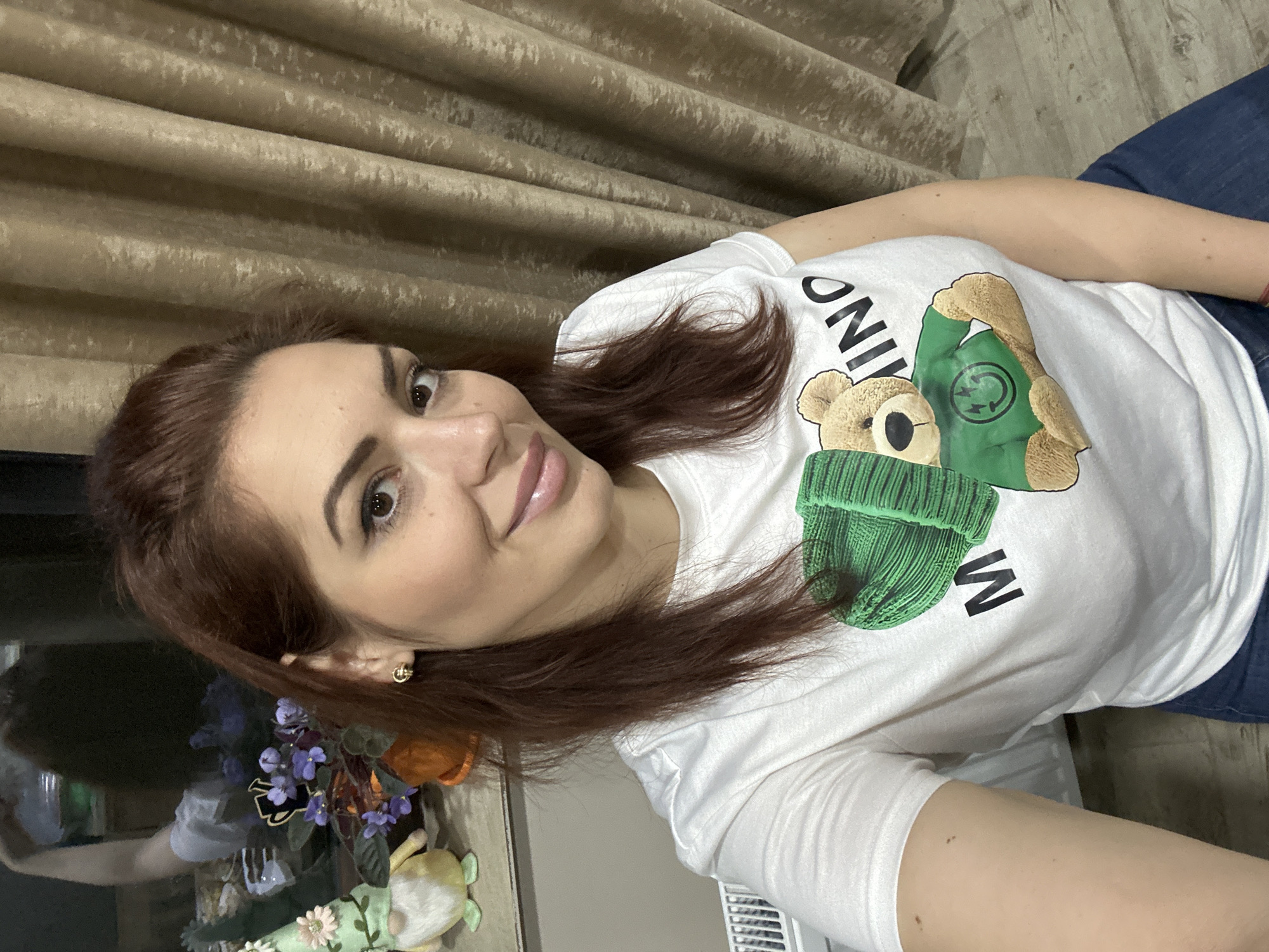 A woman with reddish-brown hair and fair skin is centered in the image, wearing a white t-shirt with a cartoon bear graphic in green. She is wearing gold earrings. The background features a neutral-toned curtain on the left and what appears to be blue denim on the right. A decorative floral arrangement is visible to the left. The location and city of Lehchevo, Bulgaria, are not discernible from the provided image.