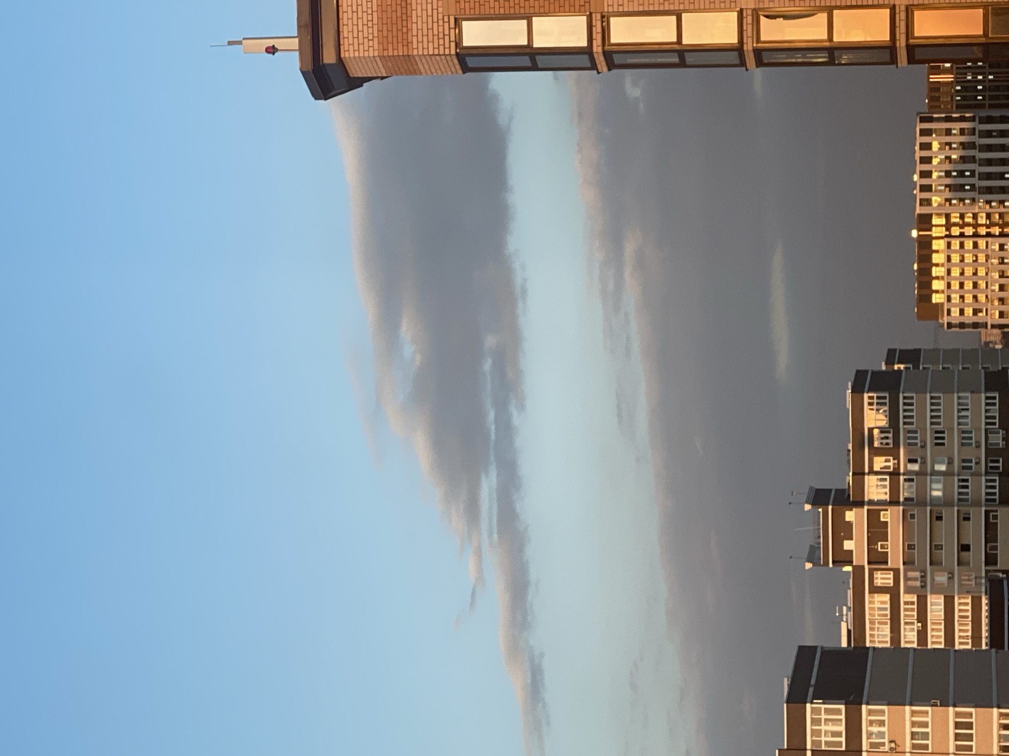 The image shows a cityscape with several multi-story buildings against a sky with clouds. The sun is setting, casting a warm golden light on the right side of the buildings. The sky is a clear blue with scattered grey clouds. In the foreground, three apartment buildings are visible. The building on the far right is made of brick and has a pointed roof with a small antenna and a red light. The building in the center is tall and modern, with many windows reflecting the golden light. The building on the left is also a modern apartment building, with a dark facade and lighter colored sections. The overall scene depicts a residential area in a city during sunset.
