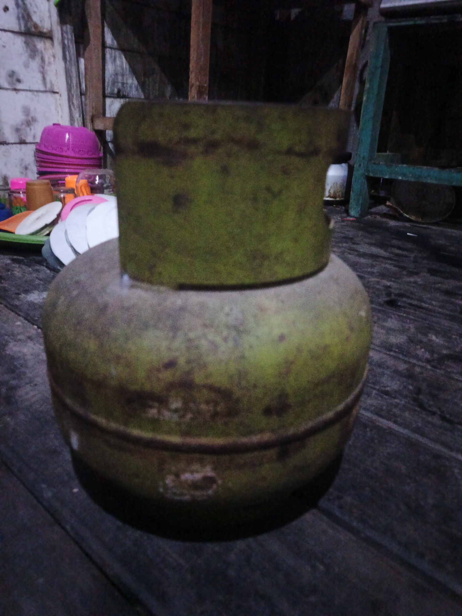 The image shows a close-up of a propane tank on a wooden floor. The tank is green and appears weathered. Behind the tank, there are some stacked pink plastic bowls, a green metal structure, and a wooden wall. The scene is dimly lit and appears to be in an outdoor or semi-outdoor setting. The specific location in Sinar Bulan, Indonesia is not determinable from the image.