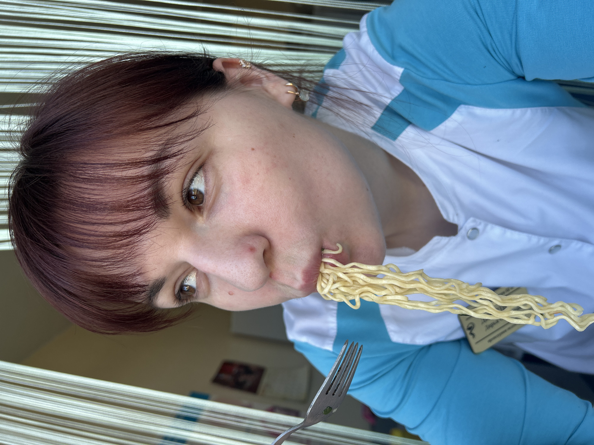 A person with dark hair, wearing a white shirt with blue sleeves, is eating noodles with a fork. The person has multiple piercings on their ear. The background includes vertical striped curtains, a wall with pictures, and a blurred interior. It is difficult to verify with certainty whether the location is Фоки, Russia, without additional contextual information.