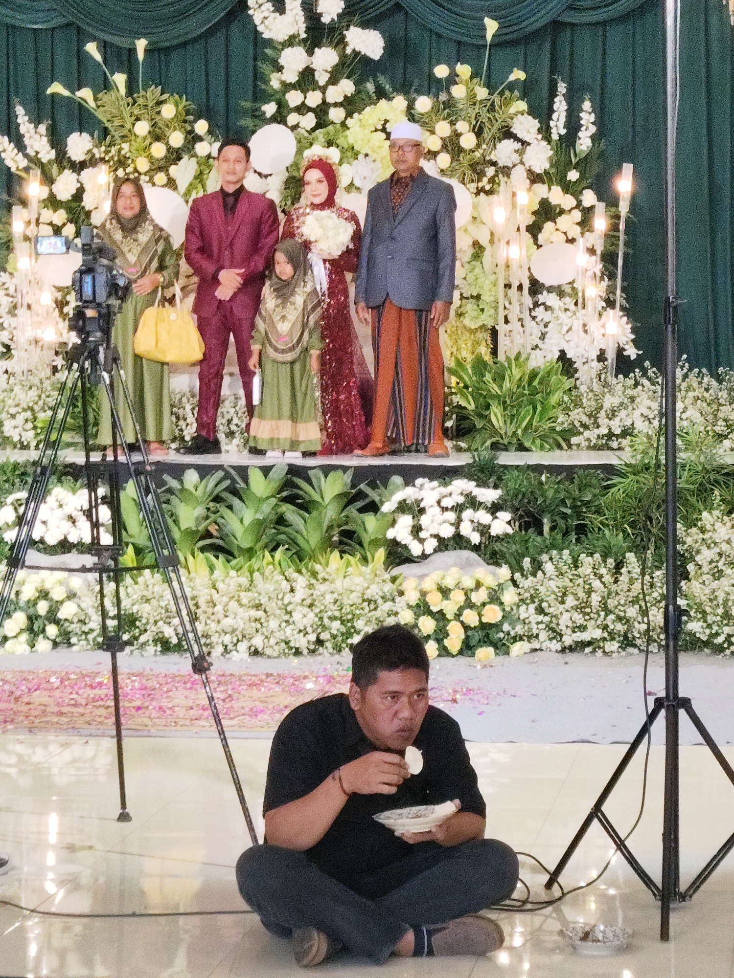 A wedding ceremony is taking place in Dolopo, Indonesia. The bride and groom are standing on a stage, surrounded by floral decorations. The bride is wearing a traditional red and gold gown, while the groom is in a dark suit. They are accompanied by two women in green dresses and a young girl in a similar outfit. A man in a suit and religious headwear stands to the right. In the foreground, a man is sitting on the floor, eating from a plate. A camera on a tripod is positioned to the left of the stage. The backdrop is a dark green curtain. The floor is tiled and reflective, with scattered flower petals. The overall scene depicts a formal event with a festive atmosphere.