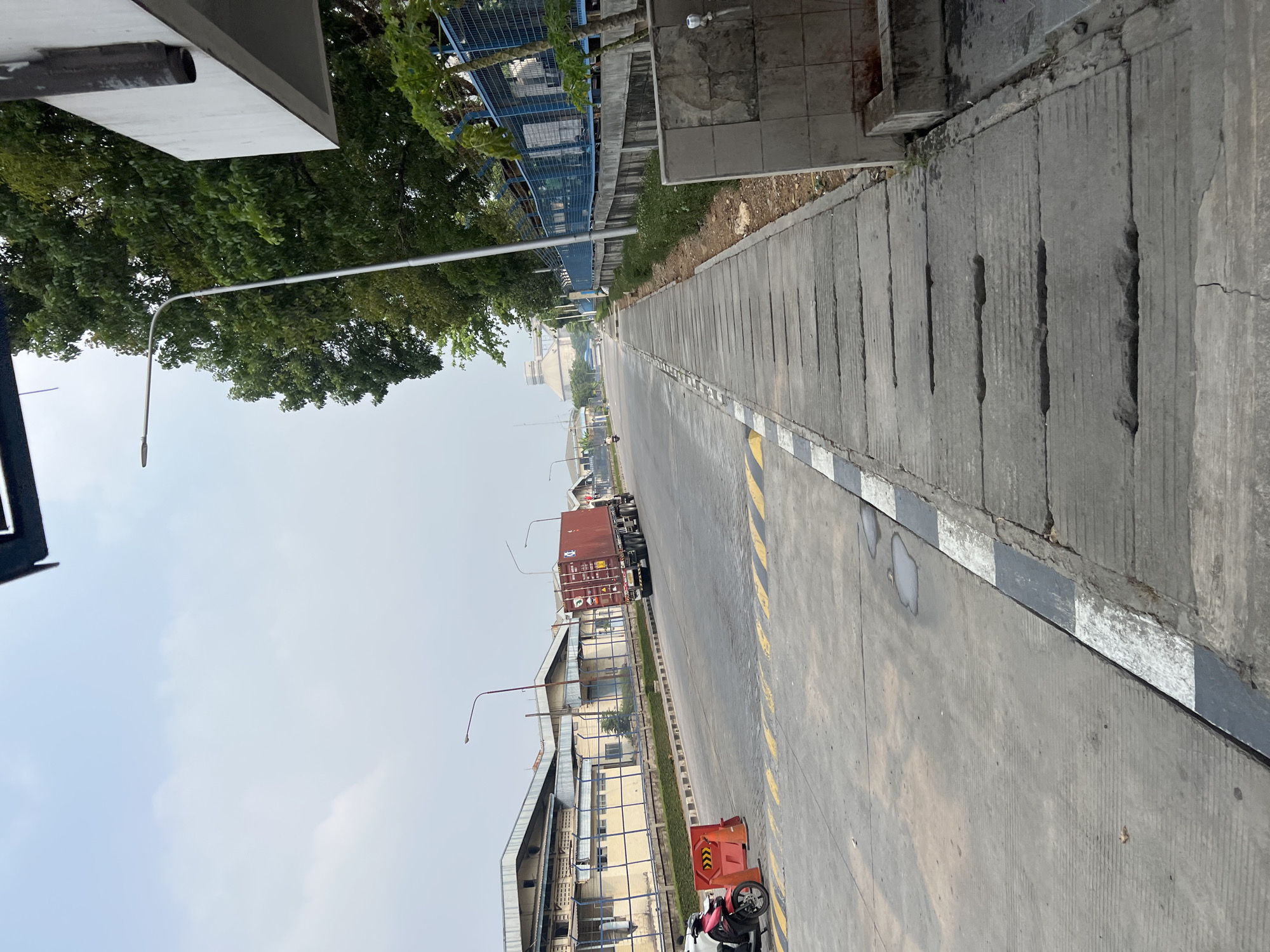 A low-angle, medium shot captures a paved road leading towards a factory complex in Klapanunggal, Indonesia, under an overcast sky. On the left, a pink motorcycle is parked near a red construction barrier with orange cones. A large red container truck travels down the road on the right, with a blue metal fence and lush green trees lining the right side of the road. In the background, the low-rise buildings of the factory complex are visible, and a street lamp stands on the right side of the road. The foreground is dominated by a concrete sidewalk with grated drains. The scene appears to be during daylight hours.