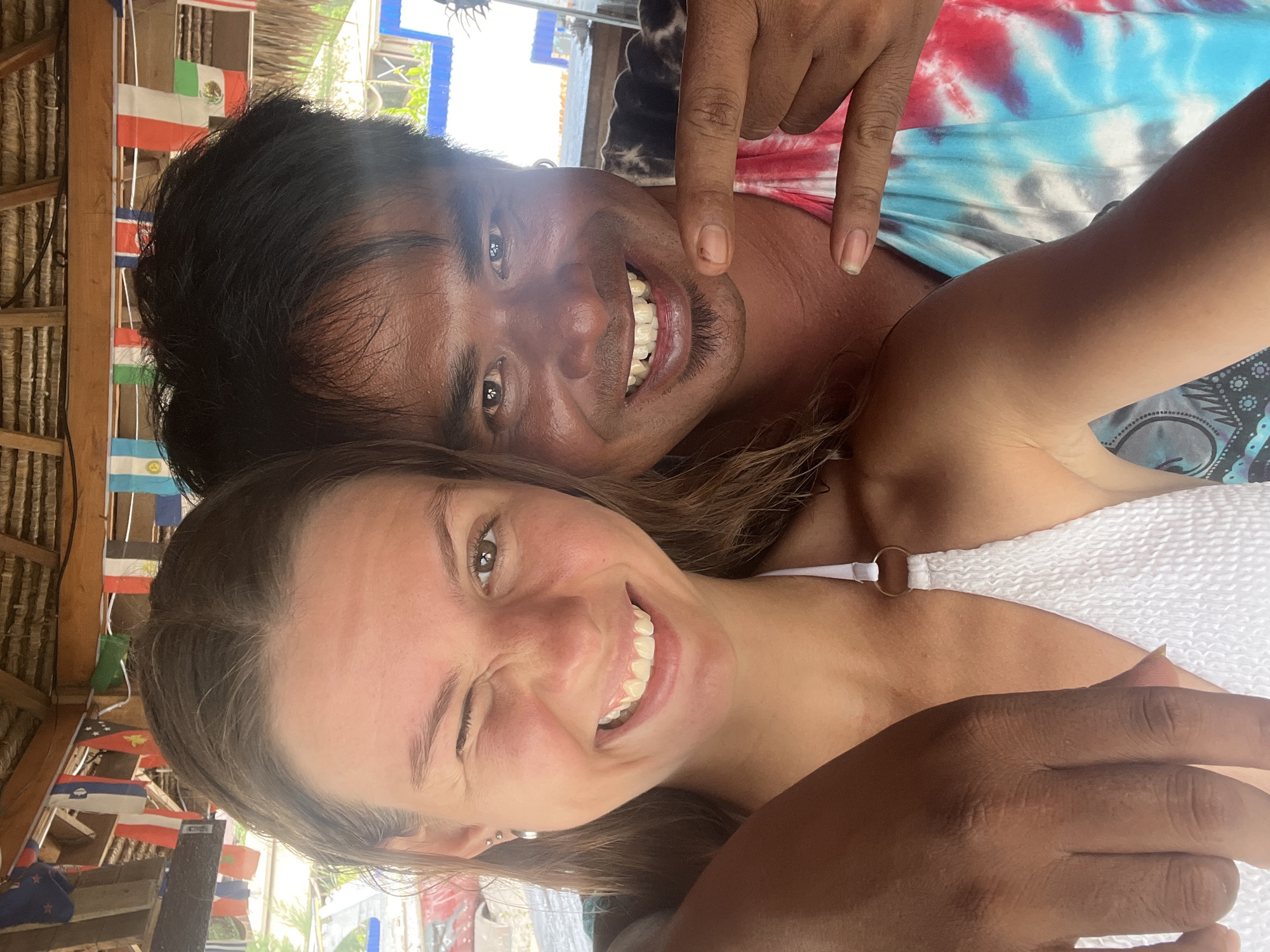 The image is a selfie featuring two individuals. The person on the left appears to be a young Caucasian woman with blonde hair, winking and smiling. She is wearing a white textured bikini top with a ring detail. The person on the right is a man with dark hair and brown skin, smiling broadly. He is wearing a tie-dye shirt. The man's right hand is in the foreground, with his index and middle fingers extended. They are both positioned closely together, suggesting a shared moment.
The background shows a thatched roof structure with numerous small flags strung across it, representing various countries. Beyond the structure, there are glimpses of buildings and greenery, characteristic of a tropical or coastal setting. The lighting suggests it is daytime. The location is identified as Unknown, Indonesia.