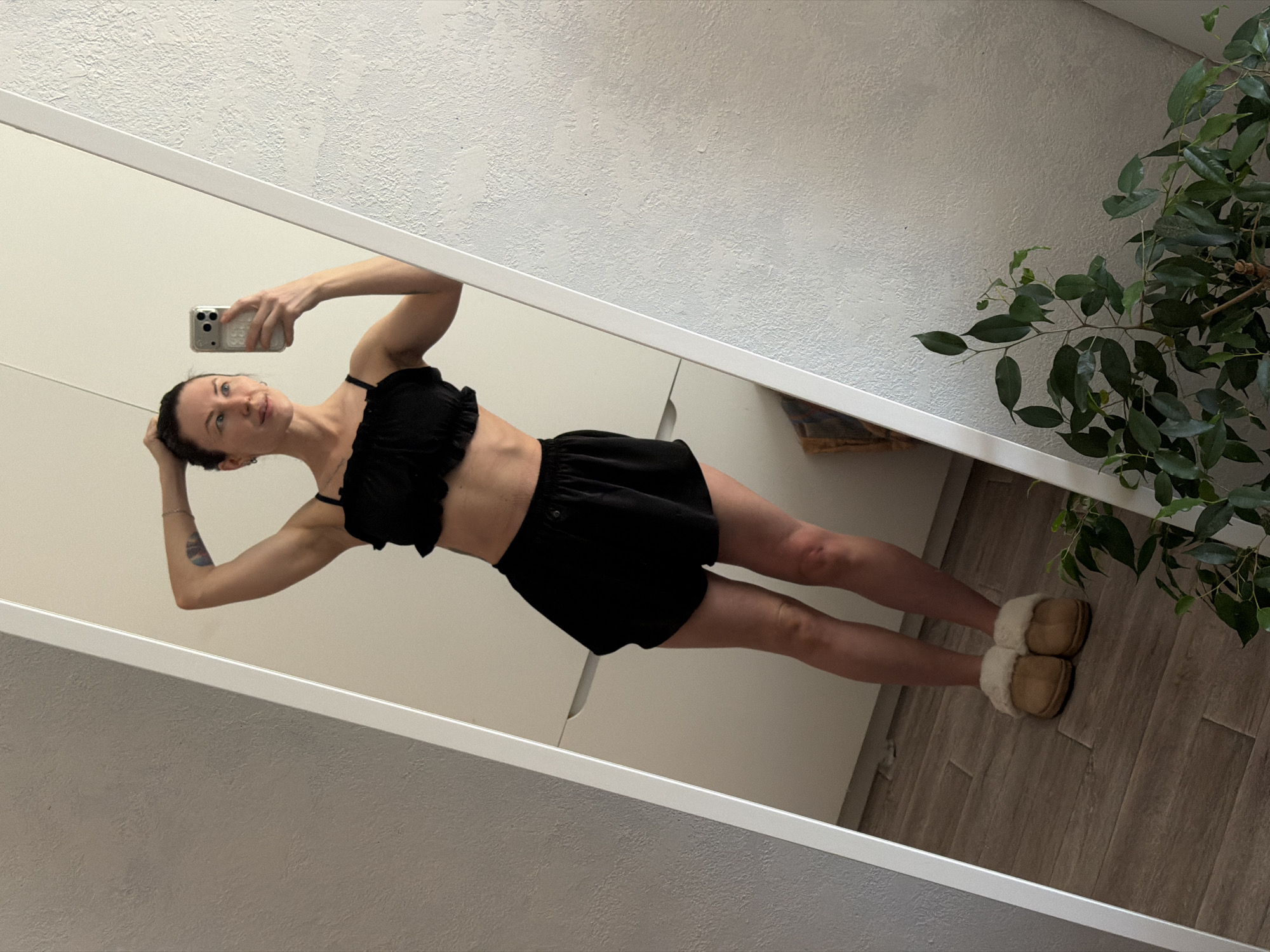A woman stands in a bedroom or dressing area, posing for a selfie in a full-length mirror. She is wearing a black ruffled crop top and black shorts, and tan slippers. The room has light-colored walls, a built-in cabinet, and a wood-look tiled floor. A potted plant is visible in the corner of the room, and the location is in the city of Perm, Russia.