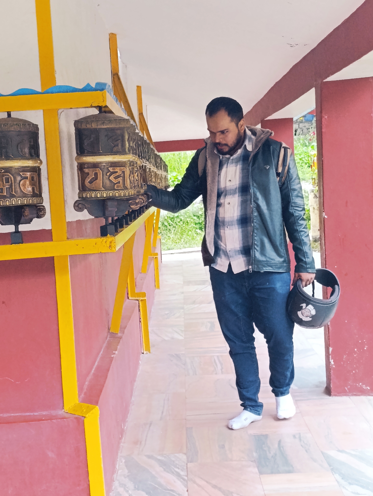 A man with a beard stands on a marble-tiled floor in front of a wall painted red and yellow. He is wearing a blue and white plaid shirt, a black leather jacket with a grey fleece lining, blue jeans, and white socks with white sneakers. He is holding a black helmet in his right hand. To his left, a row of prayer wheels, decorated with gold lettering, are mounted on a yellow structure. The man appears to be looking at the prayer wheels. The background shows greenery and a red-painted architectural element. The scene is likely at a temple or monastery in Manali, India, given the prayer wheels and the general aesthetic.