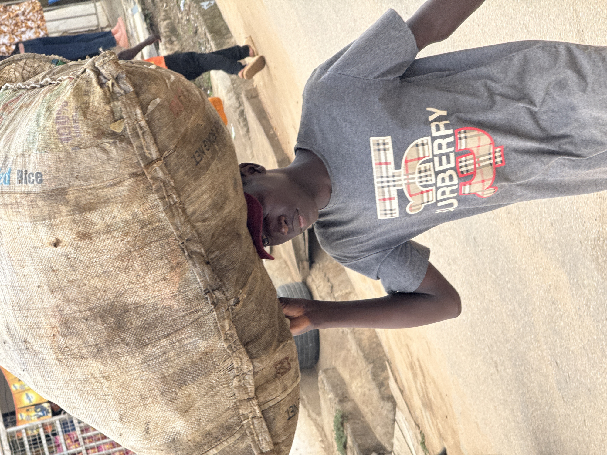 A young, dark-skinned person, likely male, is captured in an outdoor, upright pose. They are wearing a grey short-sleeved t-shirt and a dark red cap. The person is carrying a large, heavy-looking sack made of a coarse, light brown or beige woven material, resembling burlap. The sack is positioned against their head and left shoulder, obscuring most of their face except for their eyes and nose, which are turned towards the viewer. Their right arm is visible, extended down alongside the sack.
On the grey t-shirt, a distinct graphic design is visible. The word "BURBERRY" is printed vertically in white, integrated into a large stylized monogram that appears to be "TB" or "HB" filled with a red and beige plaid pattern.
Text visible on the sack includes "RICE" in blue lettering and "50KG NET" in black, suggesting the contents and weight of the sack. The sack itself appears well-used and dirty, with visible stains and some wear.
The setting appears to be an urban or semi-urban street or pathway, consistent with Abuja, Nigeria. The ground is a light-colored, possibly paved or concrete surface. In the blurry background, glimpses of other people's legs and feet are visible, as well as vague outlines of structures or goods, implying a busy environment. The lighting is bright and natural, indicating it is daytime. The person's expression is neutral, possibly showing concentration on carrying the load.