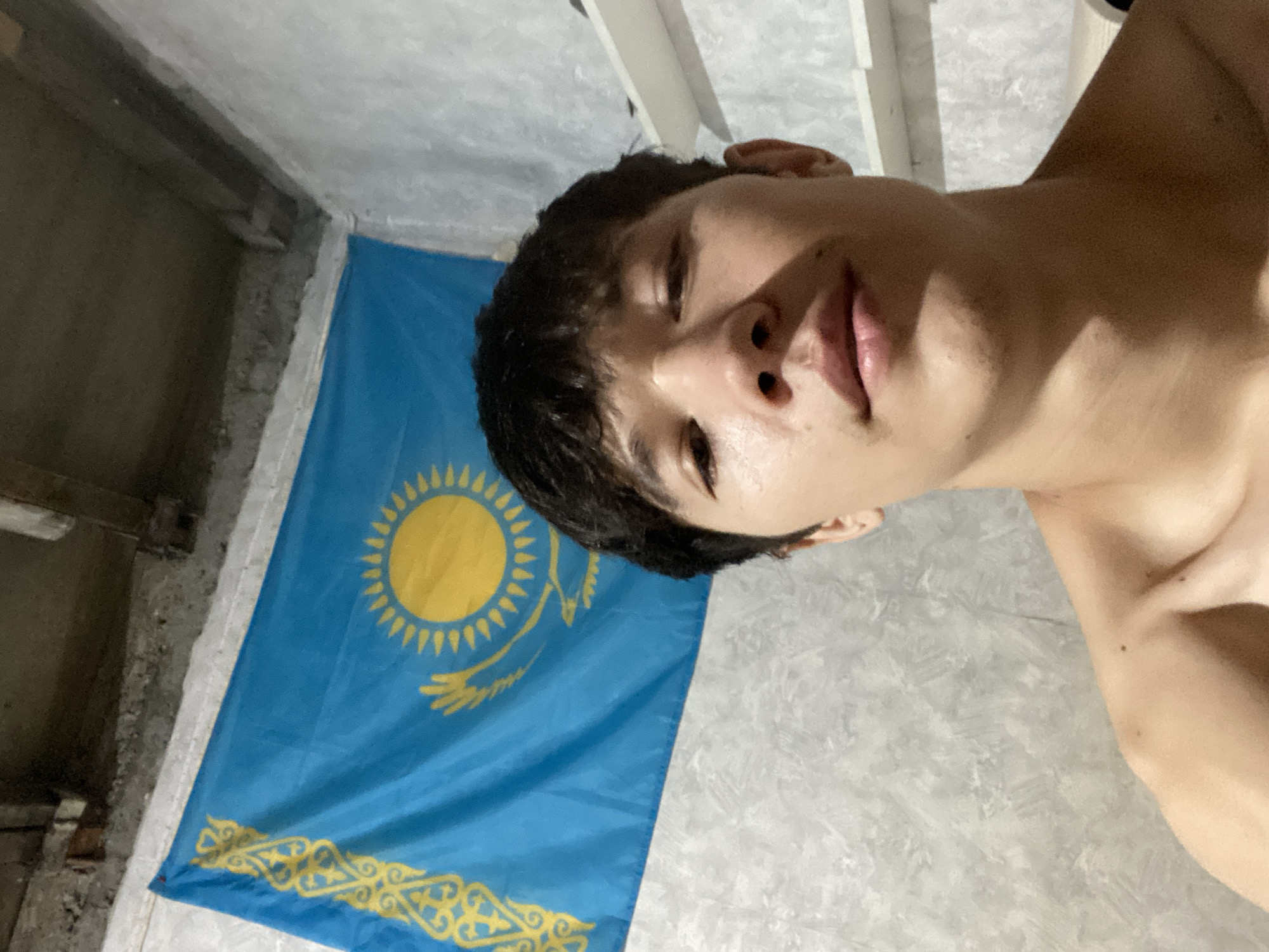 The image shows a close-up, front-facing selfie of a young man with dark, messy hair and light skin. He is shirtless, revealing his shoulders and neck. He is positioned in front of a wall, and behind him is a flag of Kazakhstan, which is blue with a yellow sun and eagle motif. The background also shows a textured grey wall and some indistinct shelving units. The lighting suggests an indoor setting. The location is not specified in the image itself, and therefore cannot be determined.