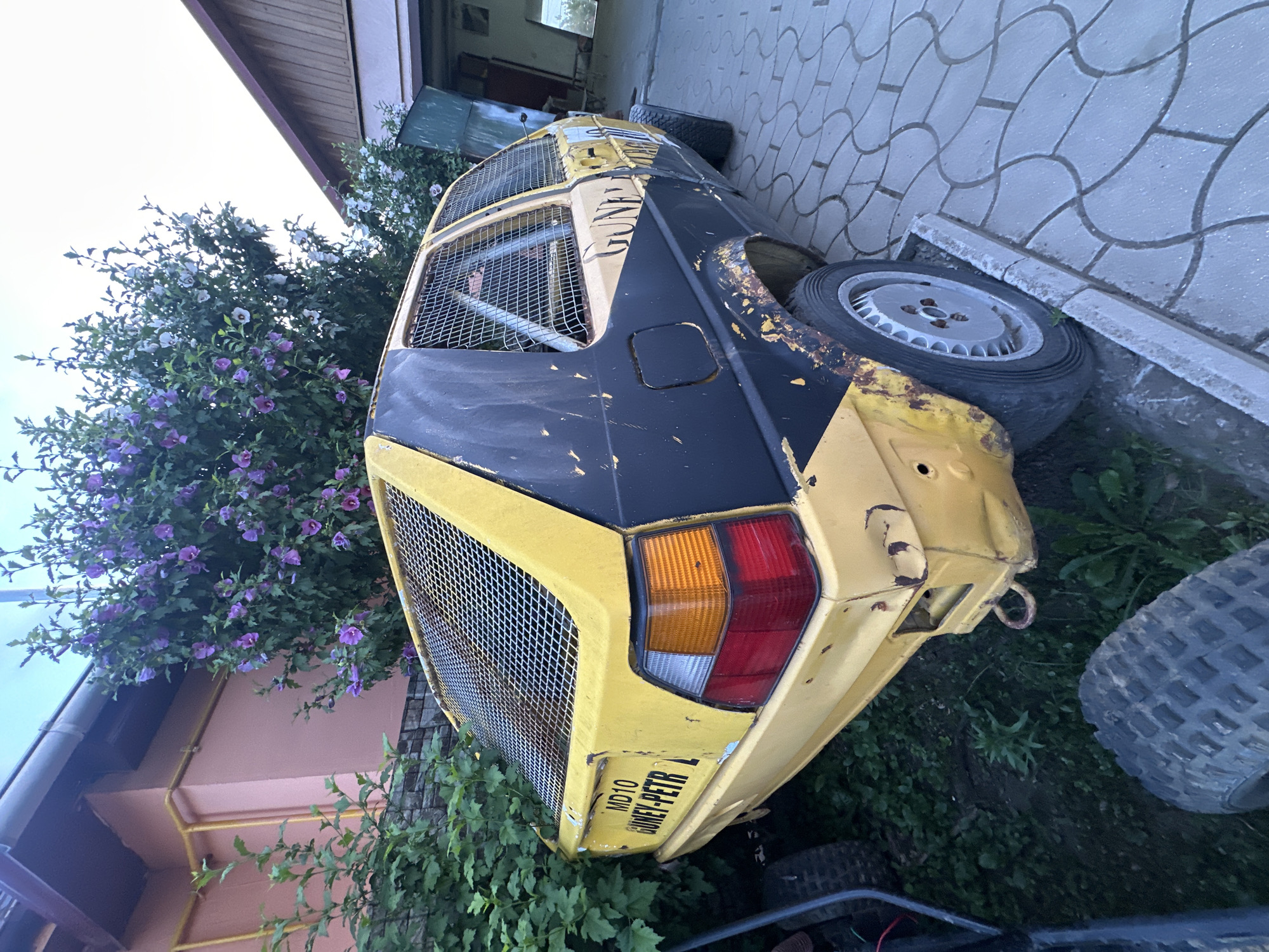 An aged, modified yellow and black Volkswagen Golf, potentially a "dune buggy" or "rally car" style, sits parked on a paver stone driveway outside a residential building with a pink facade and a wooden-shingled roof. The car is heavily weathered, with visible rust and damage. Metal mesh cages reinforce the windows and rear of the vehicle. A license plate on the rear reads "MD10 GUNEY-PETR". In the background, a lush green bush with purple flowers is abundant, and a part of a building with an open door is visible. The scene is likely in Comrat, Moldova, given the license plate format.