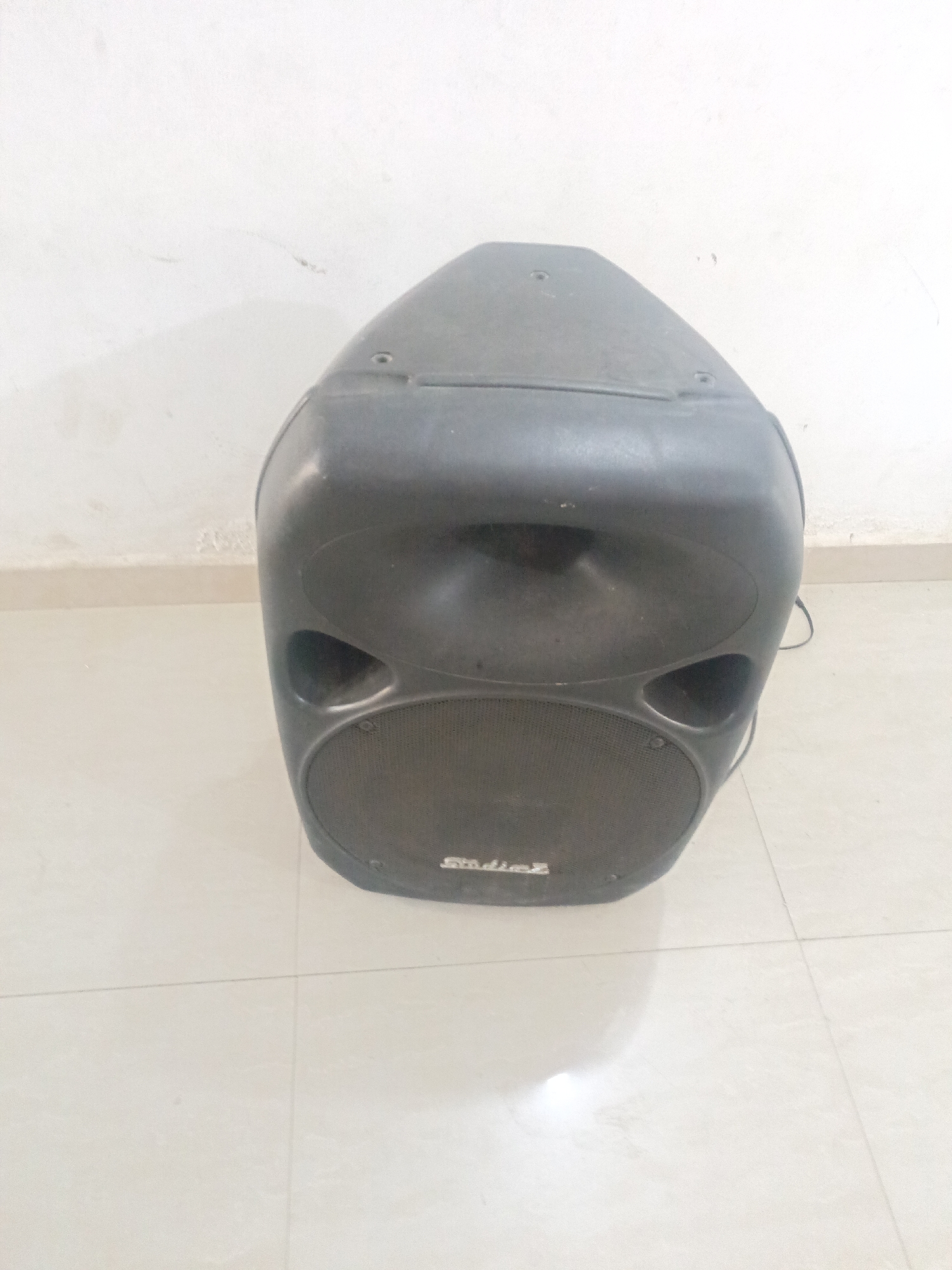 A single, used, black "Studio Z" PA speaker stands indoors on a light-colored tiled floor against a white wall. The speaker is centered in the frame and appears somewhat dusty. The environment is simple and unremarkable. The location is not discernible from the image.