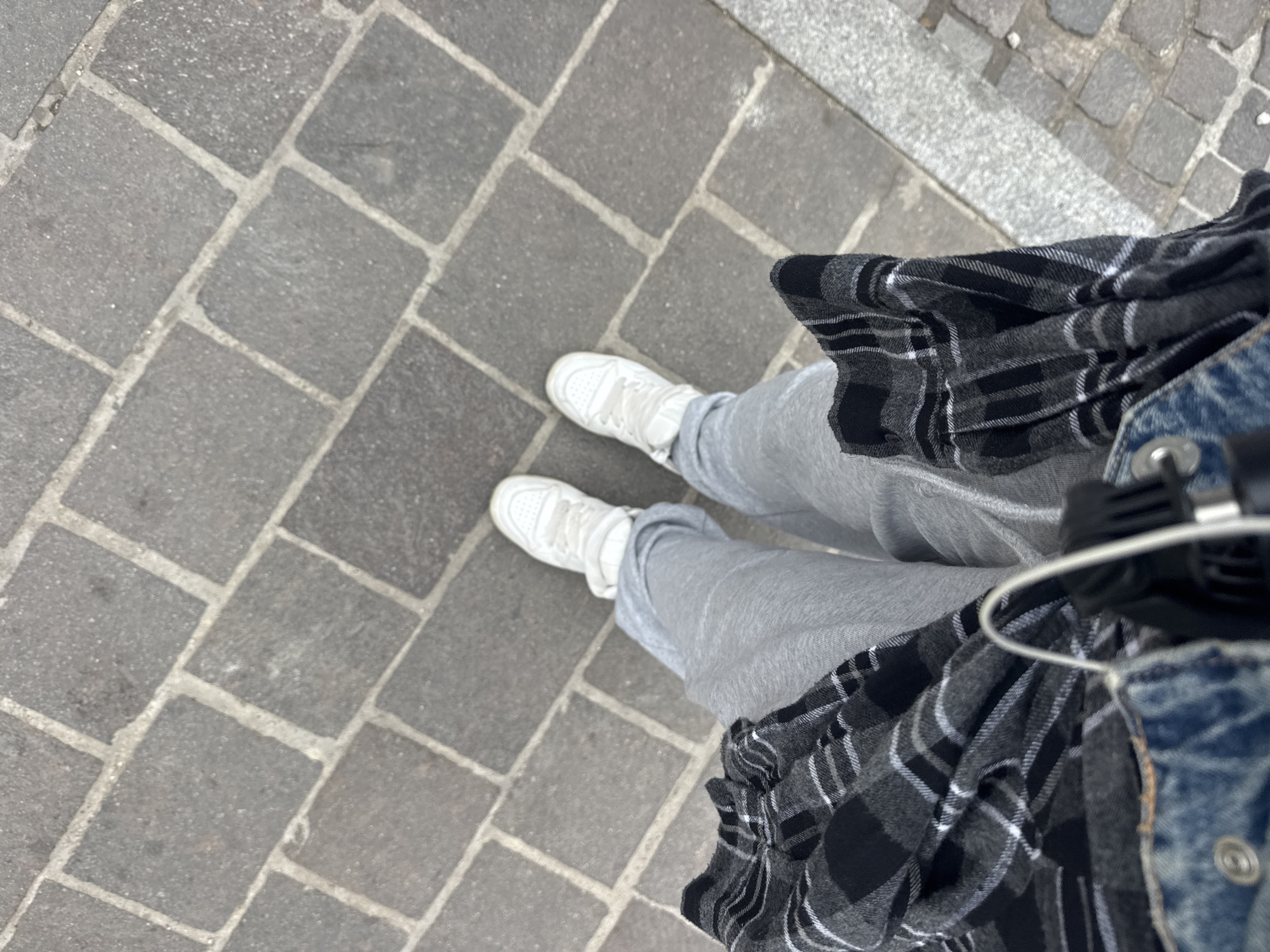 This high-angle, top-down shot captures a person's lower body and feet on a cobblestone surface in Budva, Montenegro. The individual is wearing white sneakers, light grey sweatpants or track bottoms, and a black and white plaid flannel shirt. A portion of a blue denim jacket is visible on the right side, along with a white earphone wire leading to a black earbud or component. The ground is paved with irregularly shaped grey cobblestones, clearly defined by lighter grey grout lines, indicating an outdoor urban setting like a street or sidewalk. The scene is illuminated by daylight, but the specific time of day or weather conditions are not further discernible. The person appears to be standing or walking. There is no visible text or identifiable emotional cues.