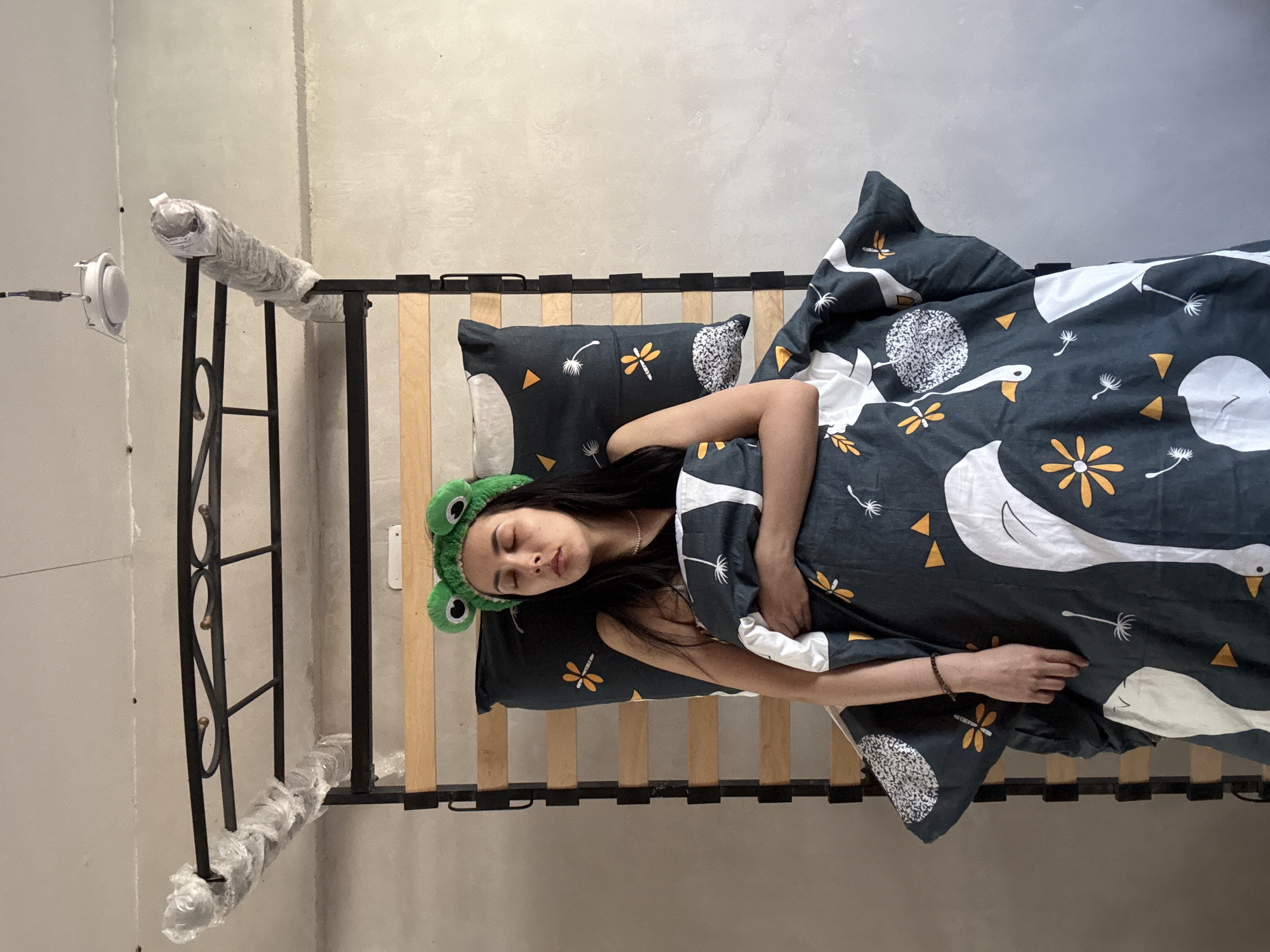 A woman is sleeping on a bed. She is wearing a green frog headband. The bed has a black metal frame and wooden slats, with some of the frame wrapped in plastic. The bedding consists of a pillow and duvet cover with a repeating pattern of white swans, yellow flowers, and geometric shapes on a dark background. The room has light gray walls and a white ceiling light fixture. The exact location in Votkinsk, Russia is not discernible from the image.