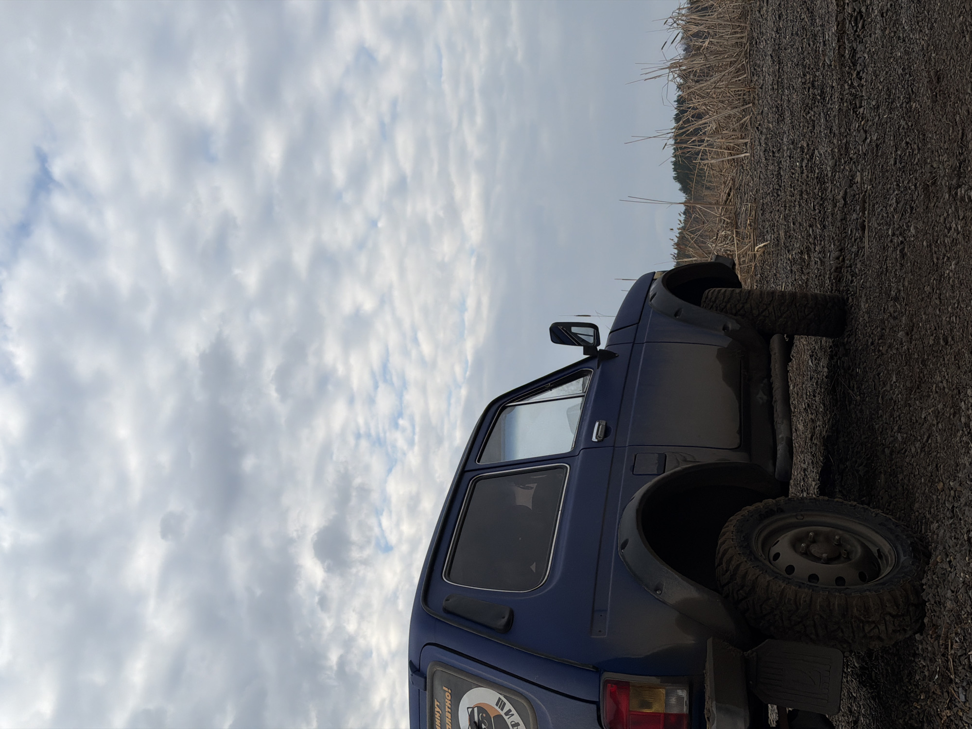 A blue Lada Niva stands on a gravelly road in Chaikovsky, Russia, against a backdrop of a cloudy sky and some reeds/grass on the edge of the road. The vehicle appears to be off-roading. The rear tire is muddy.