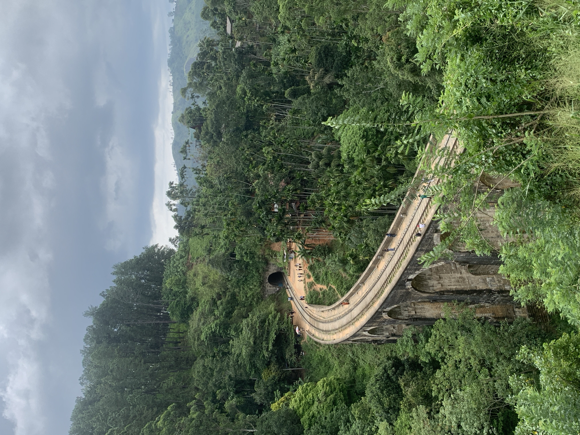The image captures the iconic Nine Arch Bridge in Ella, Sri Lanka. The bridge, constructed with stone and brick arches, gracefully curves through a lush, dense jungle landscape. Railway tracks run along the top of the bridge, and several people can be seen walking on and around it. In the background, a tunnel is visible, with more people gathered near its entrance. The scene is bathed in the soft light of an overcast sky, with the surrounding hills covered in a thick canopy of trees. The vegetation is predominantly green, with various shades and textures contributing to the verdant atmosphere.