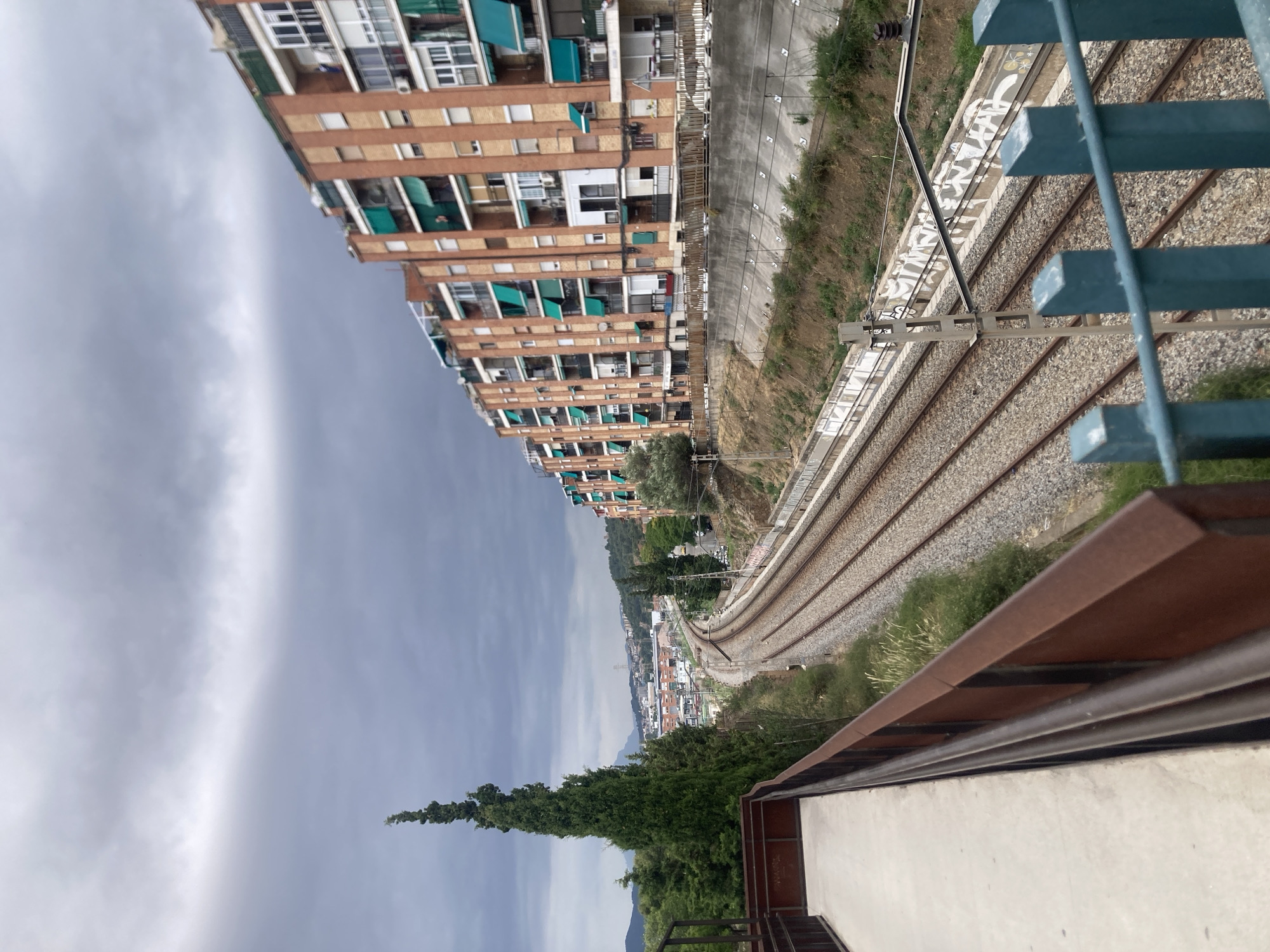 This image, taken in Sant Feliu de Llobregat, Spain, depicts a railway track curving into the distance, bordered by a long, multi-story brick apartment building on the right. The building features numerous balconies with green awnings. On the left, a tall, slender evergreen tree stands prominently, with a concrete walkway and a rusty metal railing in the foreground. Beyond the tracks, a town with smaller buildings and some distant hills are visible under a cloudy, overcast sky. The scene appears to capture an urban landscape with residential buildings and infrastructure.