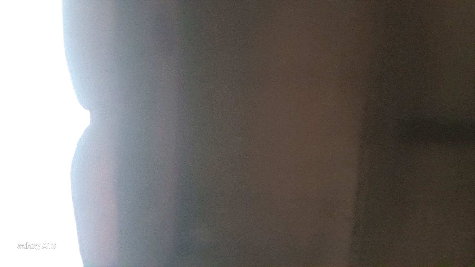 The image is a close-up, blurry shot of what appears to be a person's side profile, illuminated by a bright light source on the left. The person's body curves inward and then outward, creating a soft silhouette against a darker, textured background. The background transitions from a lighter gray on the left to a darker brown and black on the right, with visible grain and possibly subtle vertical lines. The lighting creates a gradient effect, with the left side of the subject being significantly brighter than the right. The "Galaxy A13" watermark is visible in the bottom left corner, indicating the image was captured with a Samsung Galaxy A13 smartphone. There are no discernible features to place this image in Hadipolo, Indonesia, or any specific urban environment.