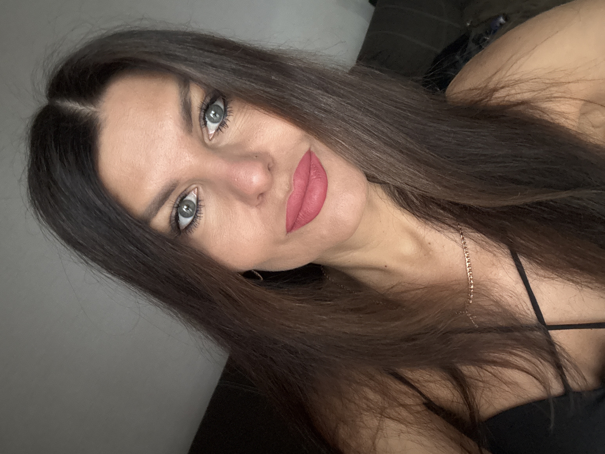 A medium-shot, head-and-shoulders selfie features a woman with long, dark brown hair and striking blue-green eyes. She is wearing a black top with thin straps and a delicate gold chain necklace. Her lips are painted a vibrant pink, and she has a small beauty mark below her nose. The background is dimly lit, with a hint of a gray wall and darker, indistinct shapes suggesting indoor furniture. The lighting casts subtle shadows, highlighting the contours of her face. The image appears to be taken in a residential setting.