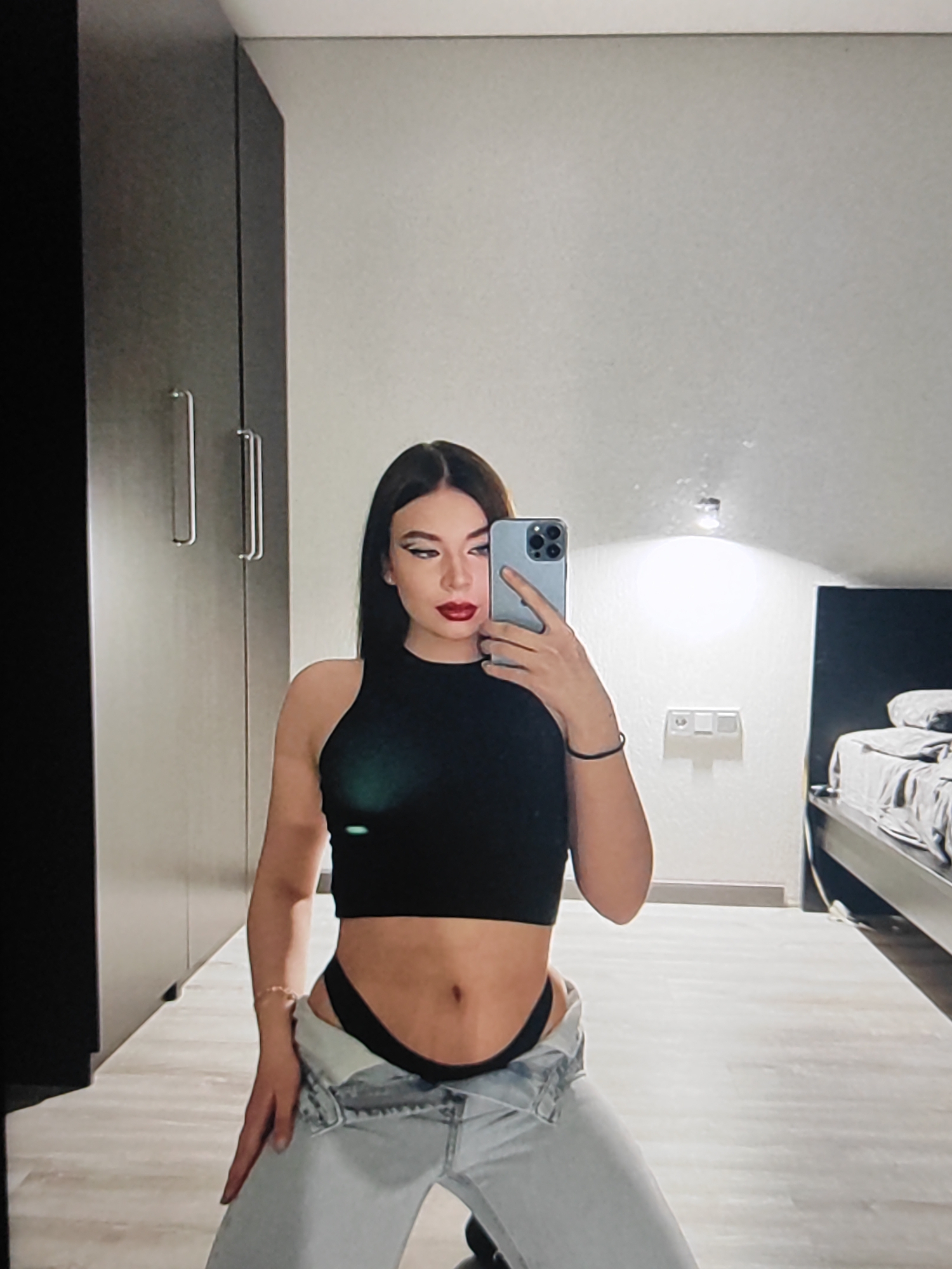 In a bedroom setting, a young woman is captured in a mirror selfie. She is wearing a black crop top and light-wash, low-rise jeans. Her makeup is bold, with winged eyeliner and red lipstick. A black bracelet adorns her wrist, and a beaded bracelet is visible on her other arm. She is holding a silver smartphone in her right hand, angled to take the photo. The background shows a tall, dark wardrobe on the left, and a bed with grey bedding on the right. A light-colored wall with a light fixture and a power outlet is visible behind her. The flooring is a light wood or laminate. The image appears to be taken indoors, likely during the day or with artificial lighting. The woman's pose is suggestive, with her body angled and her jeans pulled down.