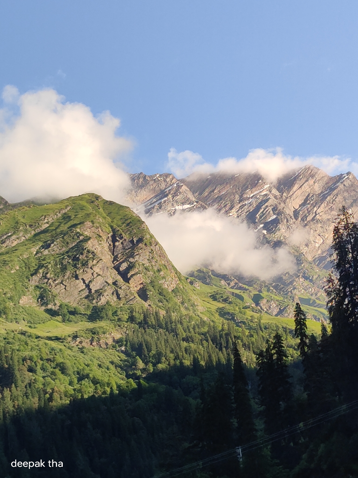 The image captures a majestic mountain landscape. In the foreground, a dense forest of pine trees covers the lower slopes, with some trees silhouetted against the brighter upper portions of the mountains. The middle ground is dominated by a steep, green mountain face, characterized by rocky outcrops and lush vegetation. In the background, towering snow-capped mountains rise majestically, partially obscured by ethereal clouds. The sky above is a clear, vibrant blue, with a few wispy white clouds adding texture. The lighting suggests it is either early morning or late afternoon, casting long shadows and highlighting the rugged terrain. The location is identified as Burwa, India. A watermark at the bottom left reads "deepak tha".
