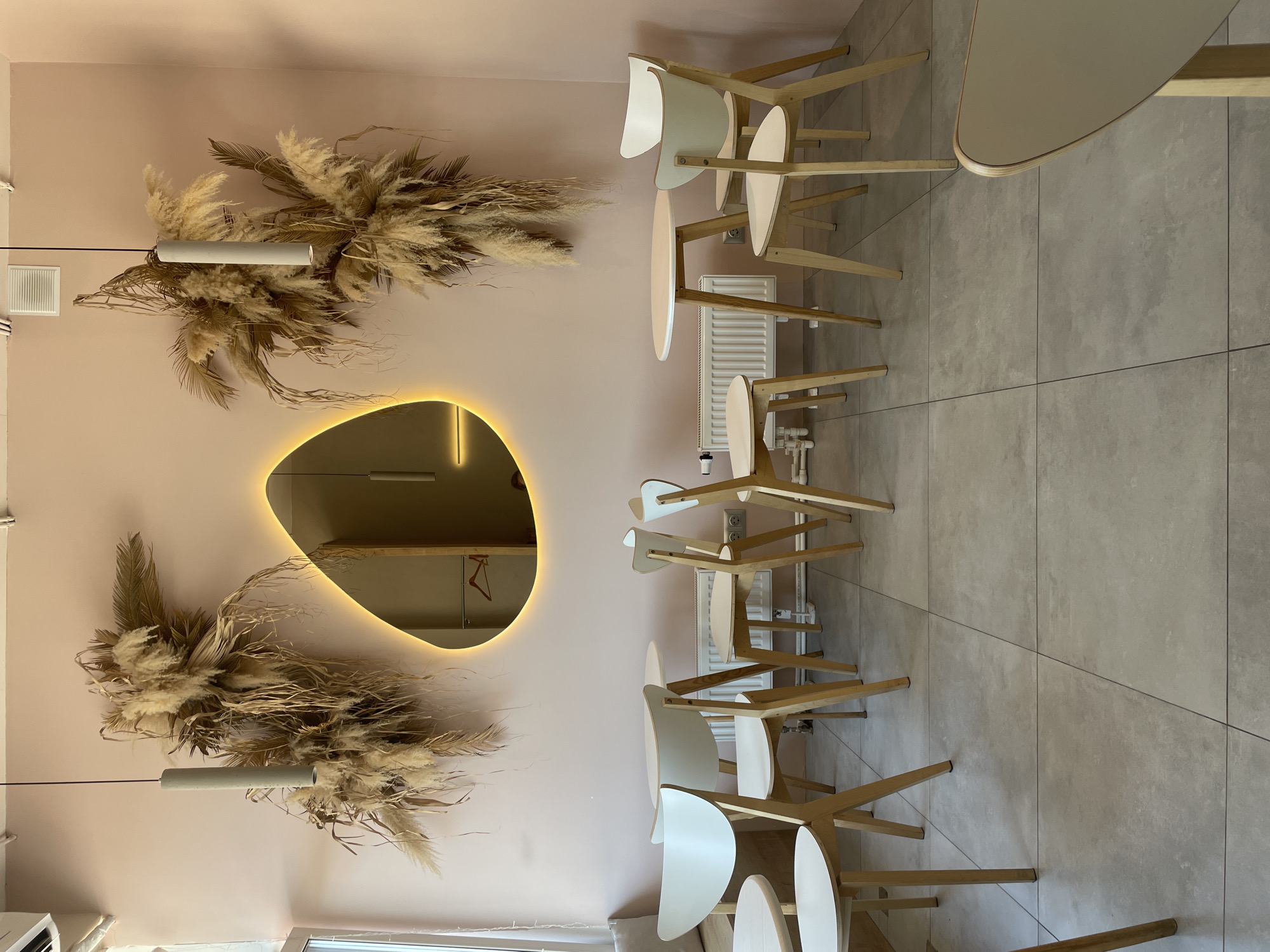 This image captures the interior of a modern, minimalist cafe or dining area. The scene is dominated by a pale pink wall on the left, serving as a decorative feature. Two large, voluminous arrangements of dried pampas grass and palm fronds are affixed to this wall, one in the upper section and one in the lower section. Each floral arrangement is accompanied by a slender, cylindrical pendant light hanging vertically.
Between these two decorations, a large, irregularly shaped mirror is mounted on the wall, featuring a warm, glowing backlighting that outlines its unique form. The mirror's reflection offers a partial view of the opposite side of the room, including what appears to be a clothes hanger. Above the upper floral display, a white rectangular air vent is visible near the ceiling.
To the right of the pink wall, a series of light-colored wooden chairs with curved white seats and backrests are neatly arranged around small, light-toned tables. These tables have smooth, slightly oval or round tops. The seating area extends along the length of the wall. A white radiator is present below an electrical outlet on the wall, with some exposed piping near the floor.
The floor throughout the area is covered with large, square, light grey tiles, creating a clean and contemporary surface. The space appears empty, with no people or visible activity taking place. The design elements suggest a calm and inviting atmosphere. No specific text is visible within the image. The location context for this setting is Vladivostok, Russia.