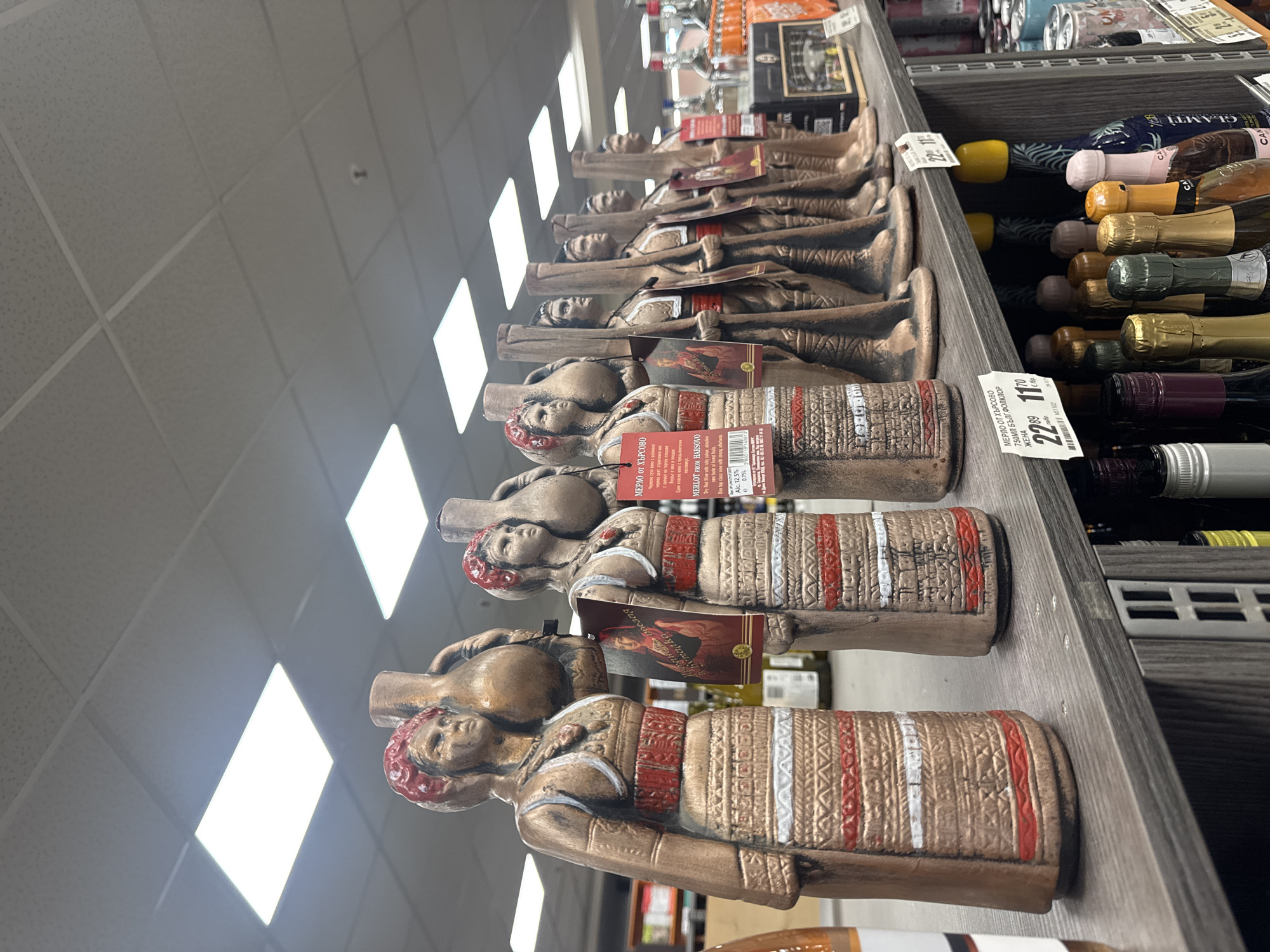 The image depicts a detailed view of a product display within a retail environment, identified as being in Plovdiv, Bulgaria, likely a supermarket or liquor store. The primary focus is a row of eight ceramic or terracotta figurines, designed as decorative wine bottles, standing upright on a dark grey laminate shelf.
From left to right, the first two figurines are female figures, each wearing a red head covering and traditional Bulgarian-style clothing with intricate red and white geometric patterns on their skirts. Each female figure holds a large, traditional jug or amphora-like vessel resting against her right shoulder. Following them are six male figurines, uniformly depicted in traditional attire including red sashes, and each carrying a long, slender object resembling a rifle or staff held upright. The figurines exhibit a rustic, unglazed ceramic appearance with painted details.
Attached to several of the figurines are product labels. One label on a female figure shows a portrait of a woman and text in Cyrillic, including "КРАСИВА РОЗИНА" (Beautiful Rozina). Other labels prominently display "МЕРЛО ОТ ХЪРСОВО" (Merlot from Harsovo), "750ML БЪЛГ. ФОЛКЛОР" (750ML Bulg. Folklore), and specify "Dry Red Wine." Price tags associated with the "Merlot from Harsovo" product are visible, indicating prices such as "22 89 лв." and "11 70 лв." (Bulgarian Lev). Another small white tag also shows "22 89" and "11 70".
Below the display shelf, a lower shelf is visible, stocked with various bottles, primarily sparkling wines and spirits, distinguishable by their diverse cap colors and bottle designs. In the background, beyond the display, the blurred outlines of other store aisles and shelves containing numerous products are discernible. The ceiling above features a typical commercial drop ceiling with square acoustic panels and multiple rectangular, brightly illuminated LED or fluorescent light panels, ensuring a well-lit environment. No individuals are present in the immediate foreground of the scene.