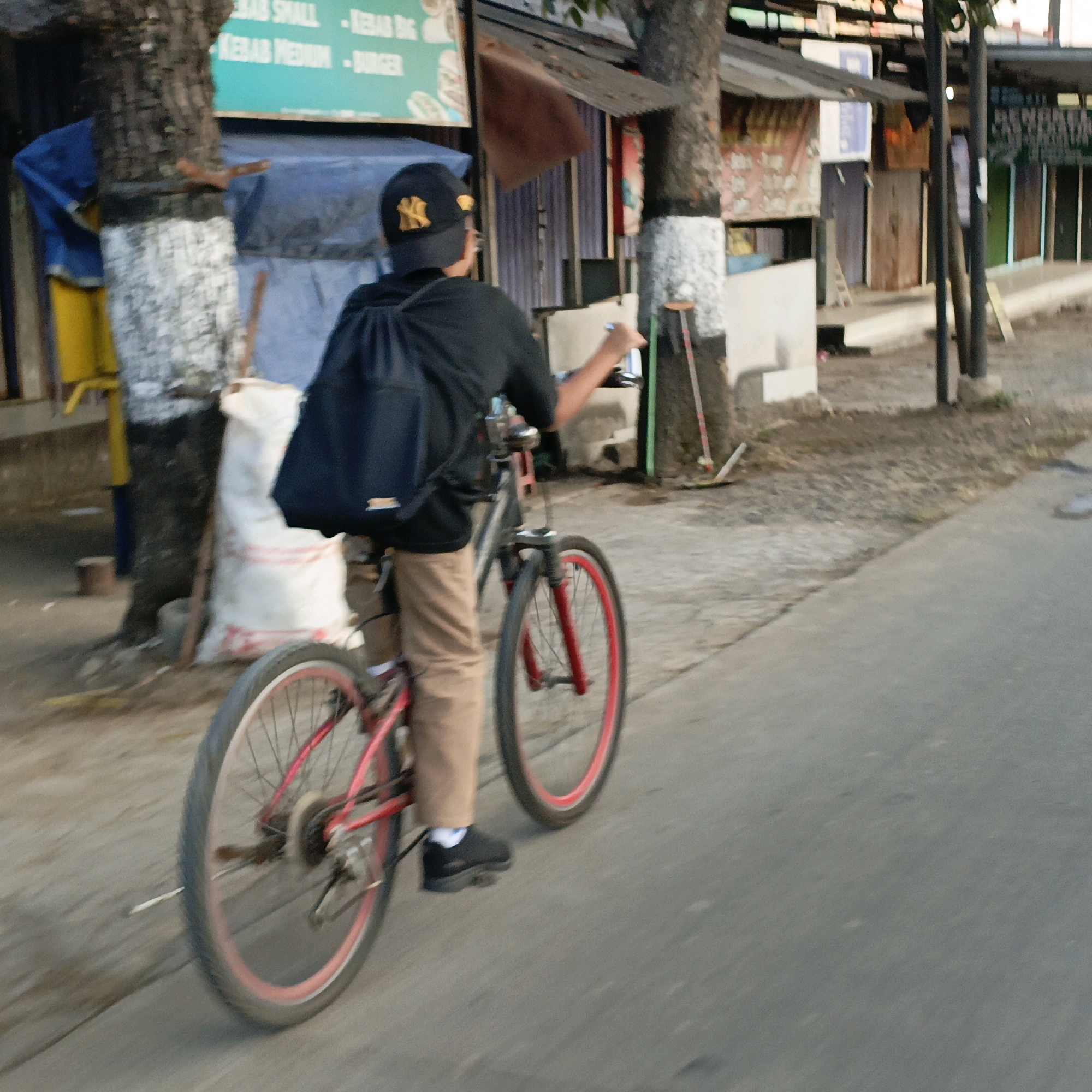 In Pabelan, Indonesia, on a paved street with a dirt shoulder, a person wearing a black t-shirt, tan pants, a black baseball cap with a yellow "NY" logo, and black shoes is riding a bicycle. The bicycle has a red frame and red rims on the wheels. The rider has a black backpack on. Behind the rider, a large white sack with red markings is leaning against a tree trunk that has white paint applied to its lower section. In the background, there are structures that appear to be small businesses or stalls, some with signage, and trees. The lighting suggests it is daytime.