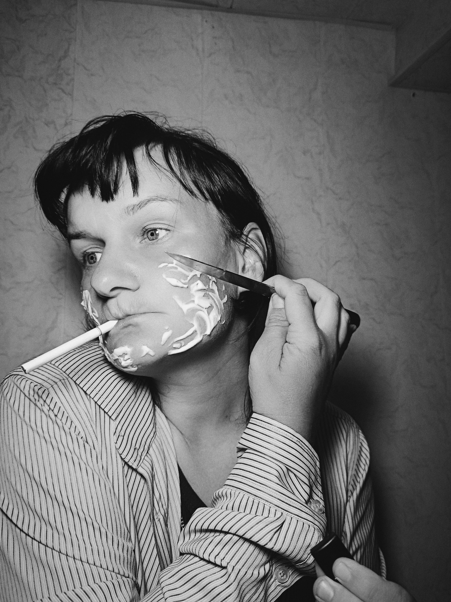 A black and white medium shot shows a person with short, dark hair in a vertically striped shirt. The person has shaving cream lathered on their face and holds a lit cigarette in their mouth. The person is also holding a razor with their right hand against their face. A small black tube is held in the lower right of the image, partially obscured by the striped shirt. The background is a grainy, textured wall. The location of Фоки, Russia is not discernible in the image.