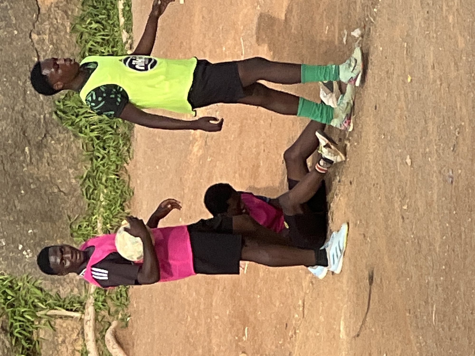 Two young men are playing soccer on a dirt field in Jos, Nigeria. One man is standing, holding a soccer ball and wearing a pink vest over a black shirt and black shorts. The other man is standing, wearing a neon green vest over a black shirt with a green pattern and black shorts. A third young man is sitting on the ground in a pink vest and black shorts. The background consists of a rocky hill with some green vegetation. The ground is covered in reddish-brown dirt and there are a few scattered white plastic objects visible. The lighting suggests it is daytime.