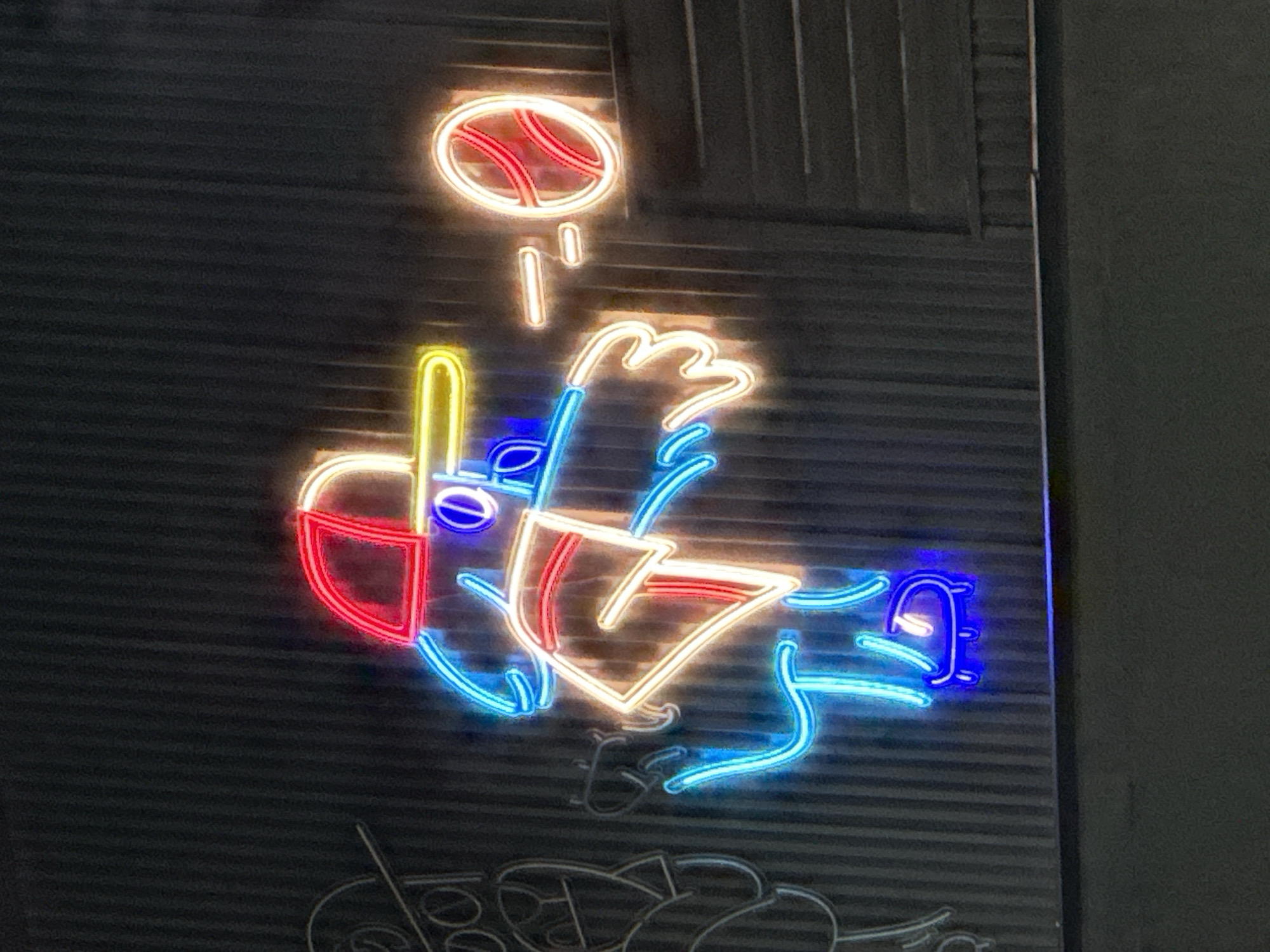 A neon sign on a wall in Unknown, Unknown depicts a baseball being pitched towards home plate. The ball is white, and the seams of the ball are depicted in red. The pitcher's hand is yellow, and the pitcher's arm is blue. The batter's helmet is red, the batter's bat is yellow, and the batter's body is white. The catcher's mitt is blue.