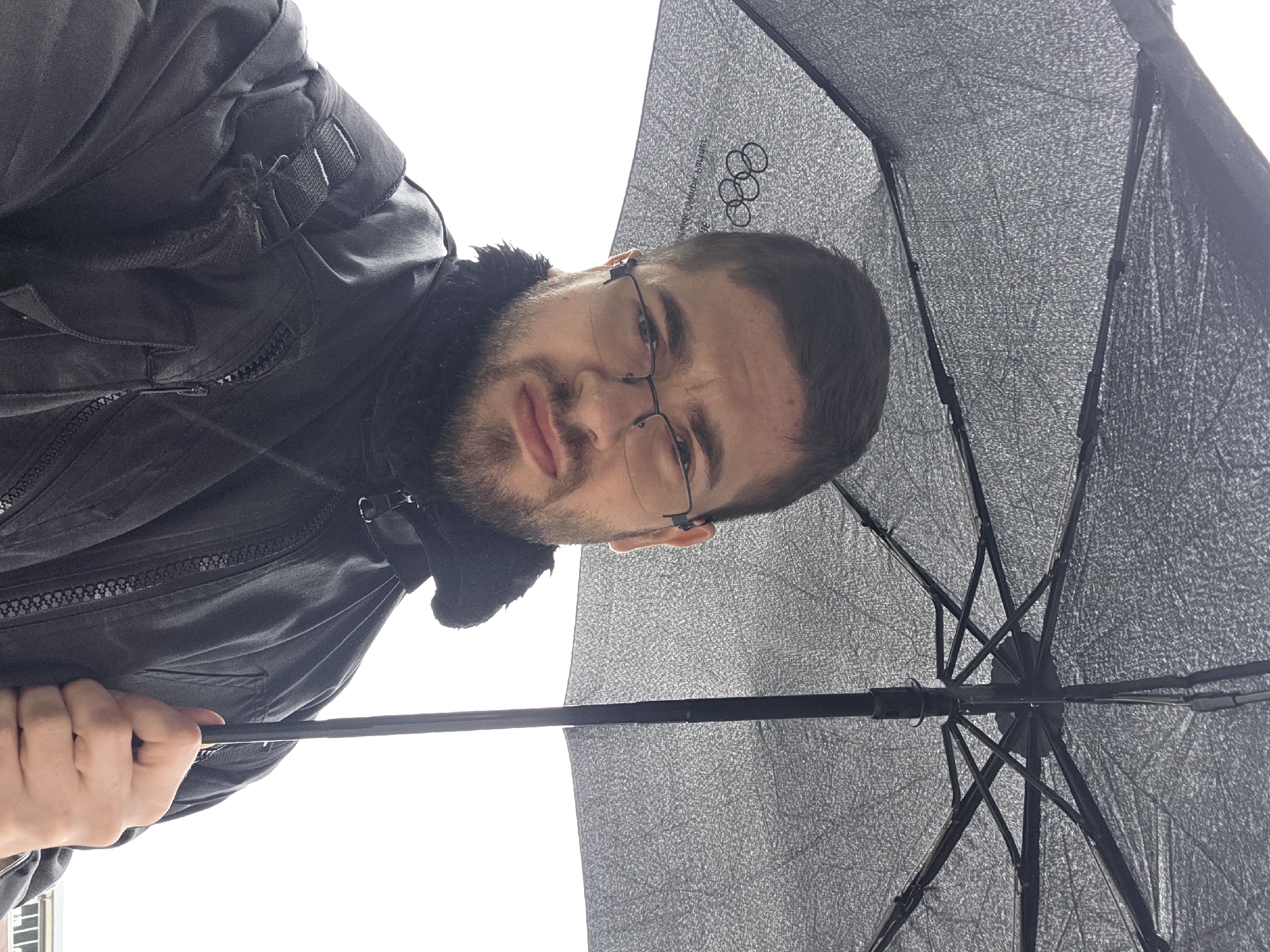 The image captures a man taking a selfie from a low-angle perspective, holding an open umbrella above him against a bright, overcast sky. The man, appearing to be young to middle-aged, has short dark hair, a neatly trimmed beard, and wears dark-rimmed glasses. He is dressed in a dark, possibly black, jacket with a fur-lined collar and prominent zippers. His expression is neutral as he looks directly at the camera. He holds the umbrella's shaft with his left hand, which is visible at the bottom left of the frame.
The umbrella's interior is a striking feature, appearing to be a textured silver or grey material, possibly for sun protection or reflection. Its black ribs and central shaft are clearly visible. Prominently displayed on the umbrella's fabric, near the man's head, is the iconic Olympic rings logo. Below the rings, reversed text is discernible, including the word "OLYMPIC" and a series of numbers, possibly dates or years such as "1964 69 70 80 90". The background is an expansive, almost pure white sky, indicating a heavily overcast day with no discernible direct sunlight or features. The scene is outdoors, and the provided context places the location in Plovdiv, Bulgaria.