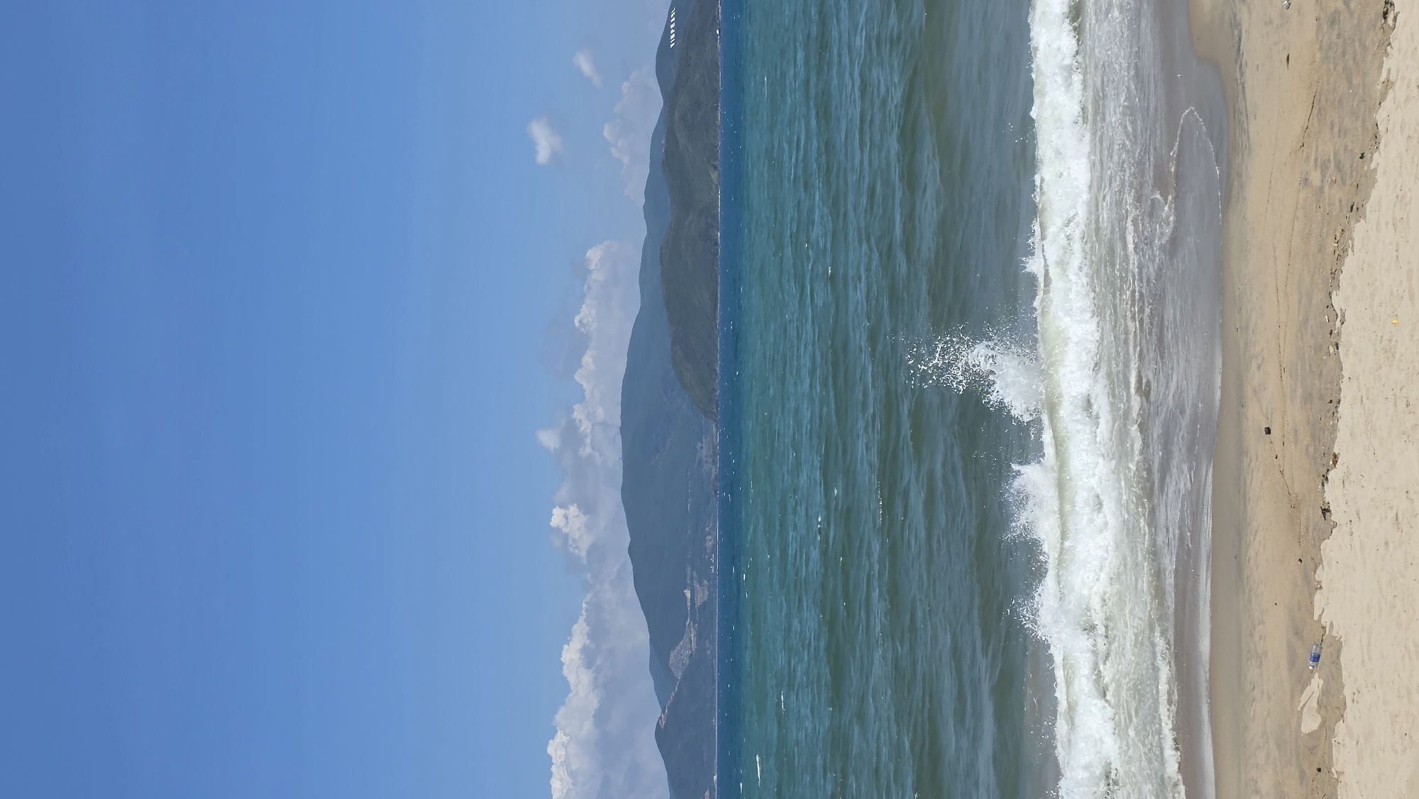 A long beach curves along the coast, bounded on one side by clear blue sky and on the other by a turbulent sea. Mountains line the horizon, partly obscured by clouds. The beach is light-colored sand, marked by the ebb and flow of waves. The location is Nha Trang Ward, Vietnam.