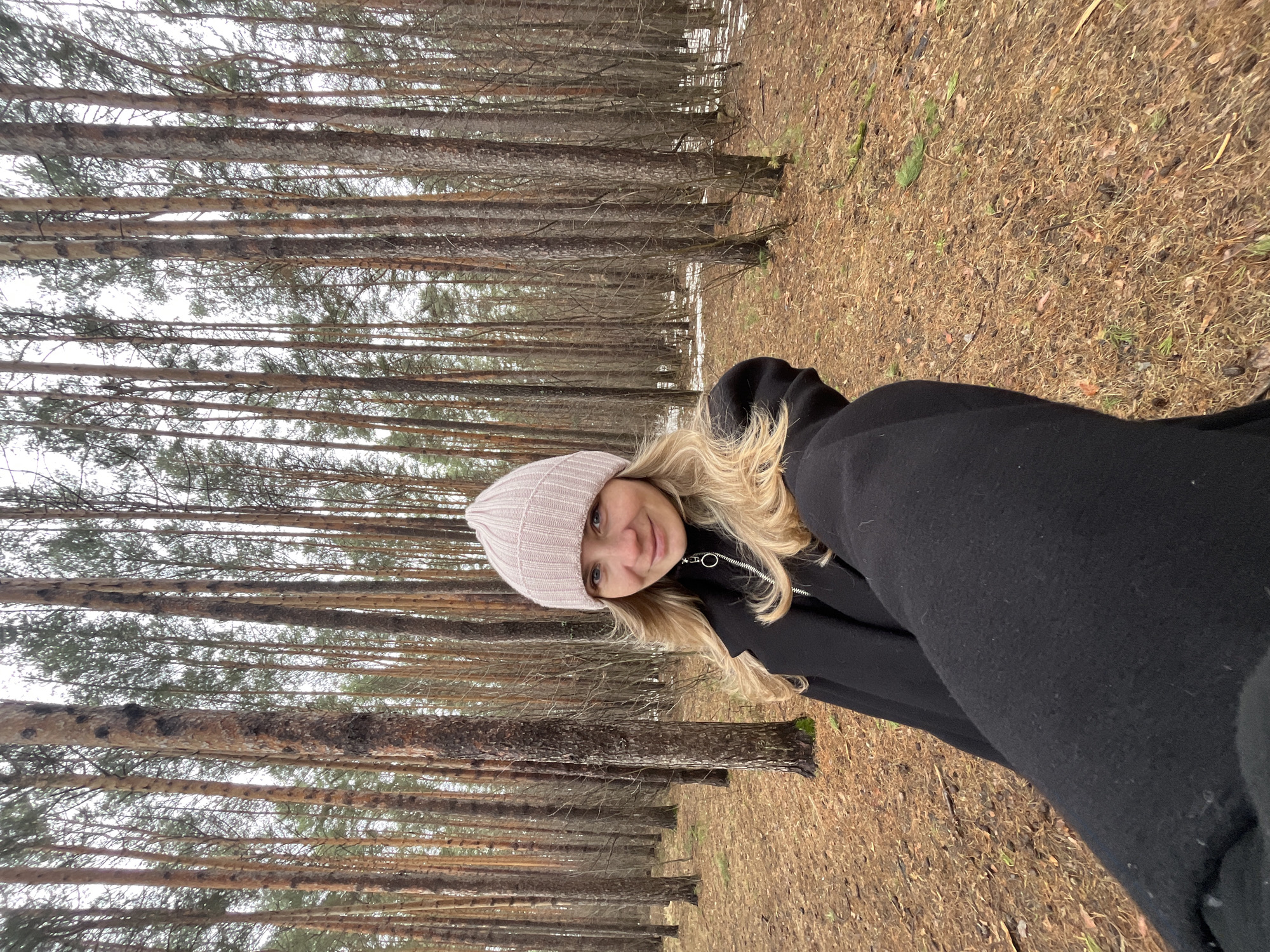 A young, blonde woman is taking a selfie in a pine forest. She is wearing a light pink beanie and a black hooded jacket. The forest consists of tall, thin pine trees with bare branches. The ground is covered in dry leaves and pine needles. There is a patch of snow visible in the background. The location is identified as a forest in the city of Chaikovsky, Russia.