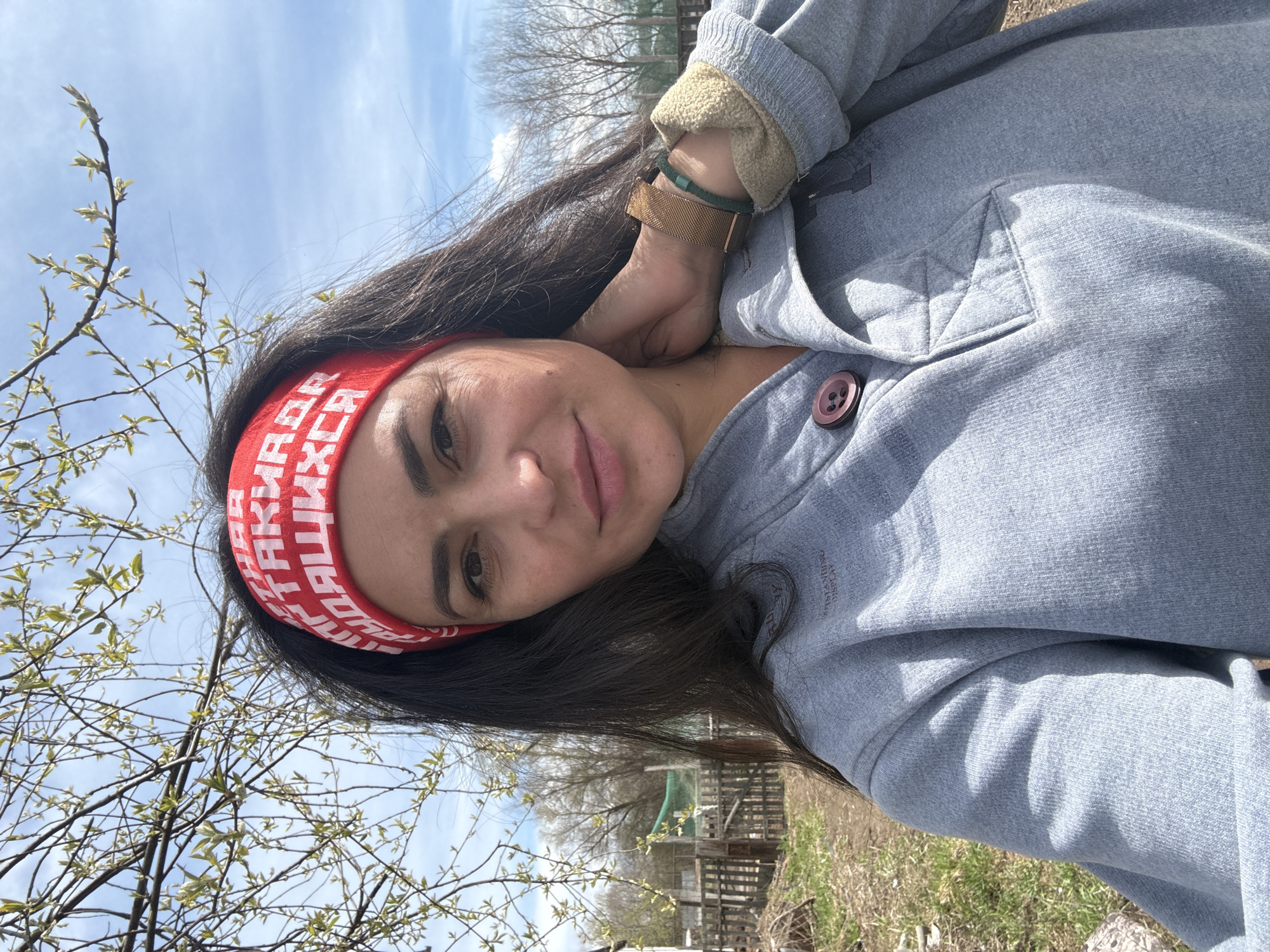 A selfie-style portrait captures a woman outdoors in Кемуль, Russia, under bright daylight conditions, suggesting spring. The woman, with long dark hair, is positioned in the foreground, smiling gently and looking directly at the camera. She wears a red headband adorned with white Cyrillic text, which appears to read "НИКТО ТАКИХ НЕ СТАВИТ ЗАДАЧ" (Nobody sets such tasks), with "ТАКИХ НЕ СТАВИТ ЗАДАЧ" clearly visible.
She is dressed in a light grey jacket with a visible brown button. The jacket features an inner lining, possibly fleece, visible at the cuff, in a light tan or cream color. On her right wrist, she wears two accessories: a gold-toned mesh watch and a green braided bracelet. The jacket also has faint text on the shoulder, reading "OUTSTANDING DISPLAY".
The background reveals an outdoor setting with bare tree branches beginning to bud with small, light green leaves, indicating early spring. Beyond the trees, a rustic wooden fence or structure is visible, along with patches of dry grass and earth. The sky is clear blue with sparse white clouds, and strong sunlight casts some shadows, suggesting a mid-day time. The overall scene conveys a calm and pleasant outdoor moment.