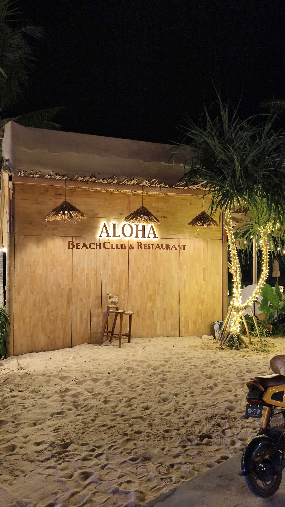 The image depicts the exterior of "ALOHA BEACH CLUB & RESTAURANT" at night. The establishment features a rustic wooden facade adorned with a prominent illuminated sign that reads "ALOHA BEACH CLUB & RESTAURANT." Three straw pendant lights hang from the ceiling, adding to the tropical ambiance. A single wooden stool stands in front of the entrance.
The scene is set on a sandy beach, with footprints visible in the sand. To the right, a palm tree is wrapped with numerous twinkling lights, creating a festive atmosphere. In the foreground, a yellow electric scooter is parked, with a license plate displaying Chinese characters. The overall impression is that of a relaxed and inviting evening destination. The time of day is clearly night, and the absence of any visible weather elements suggests calm conditions. The location context of Bac Nha Trang Ward, Vietnam, further enhances the understanding of this beachside establishment. No specific activities or events are clearly depicted, but the setting suggests a place for dining and leisure.