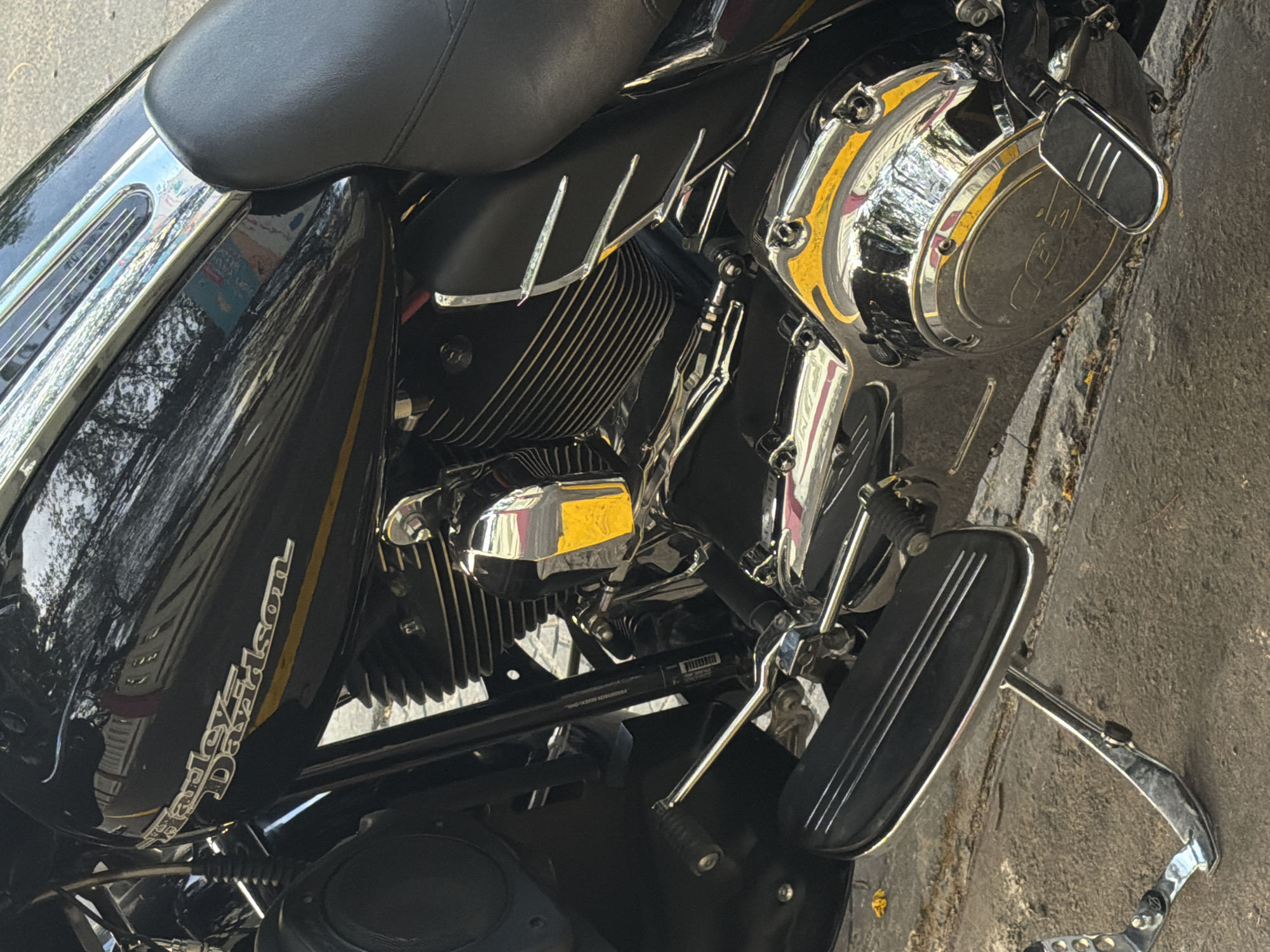 The image shows a close-up of a black Harley-Davidson motorcycle. The focus is on the motorcycle's engine, including the chrome engine cover, footrest, and fuel tank with the "Harley-Davidson" logo. The motorcycle is parked on a paved surface, possibly a street or sidewalk. The location is unspecified, and no people are visible in the frame. Therefore, it is impossible to determine if it is Đà Nẵng, Vietnam.
