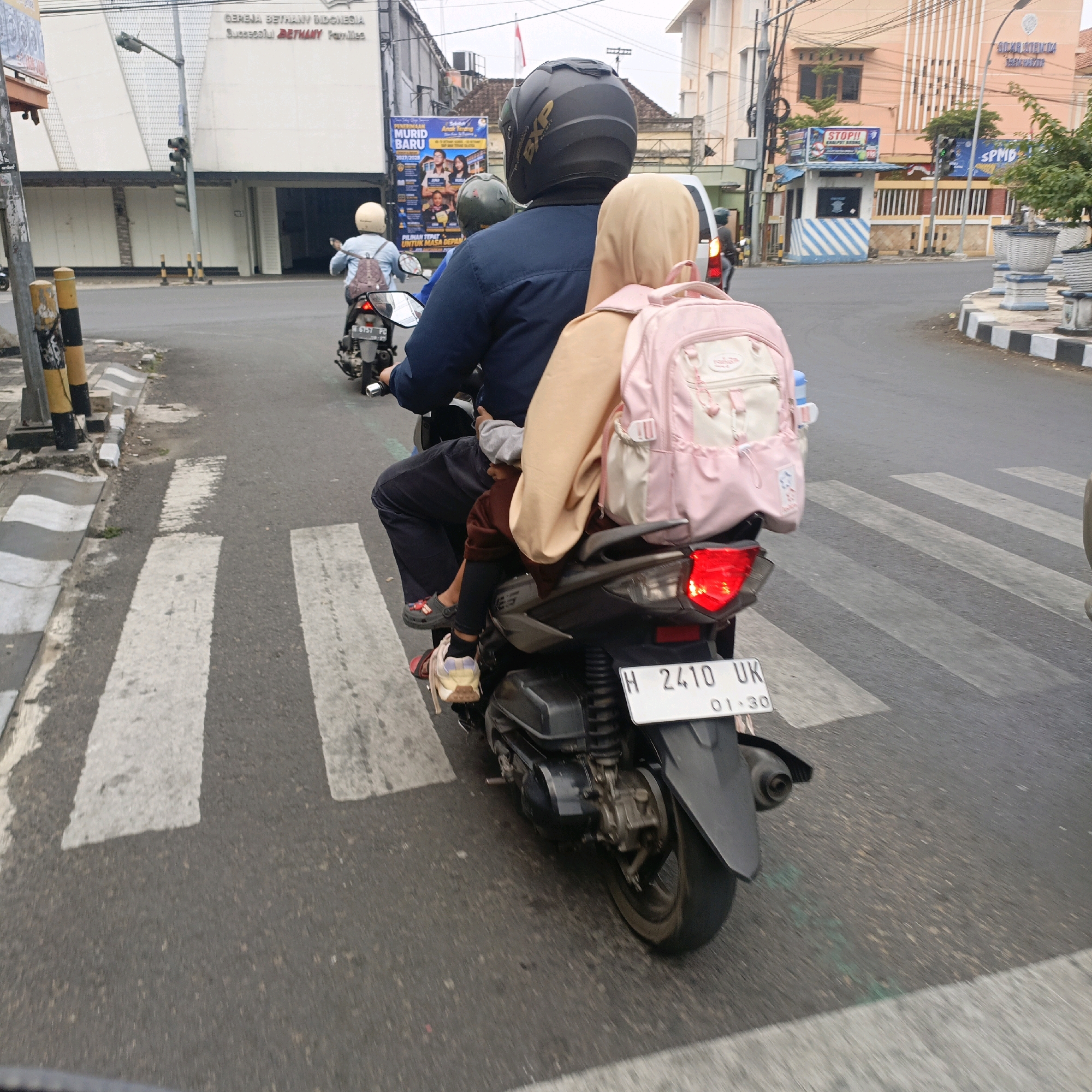 The image depicts a busy street scene in Salatiga, Indonesia, featuring three motorcycles. The main motorcycle in the foreground is a dark-colored automatic scooter with a license plate reading "H 2410 UK". It is carrying three passengers. The rider wears a dark blue long-sleeved shirt and a black helmet. Behind the rider, a woman in a tan hijab and a tan jacket is seated, also carrying a light pink backpack. A young child, possibly a girl, is seated between them. The child wears a brown long-sleeved shirt, black leggings, and a pair of dark grey sandals. The child's legs are visible, with one foot resting on the scooter's footrest. The second motorcycle, also an automatic scooter, is slightly ahead and to the left, with a rider wearing a light blue long-sleeved shirt and a light-colored helmet, also carrying a backpack. The third motorcycle is further in the distance. The road surface is asphalt with white pedestrian crossing lines. In the background, buildings are visible, including a cream-colored building with the text "GEREJA BETHANY INDONESIA" and "Successful Bethany Families" and a light orange building with "SEKOLAH STENDA TARAKANITA". Traffic lights and street poles are also present. The overall atmosphere suggests a typical day of commuting in an Indonesian city.