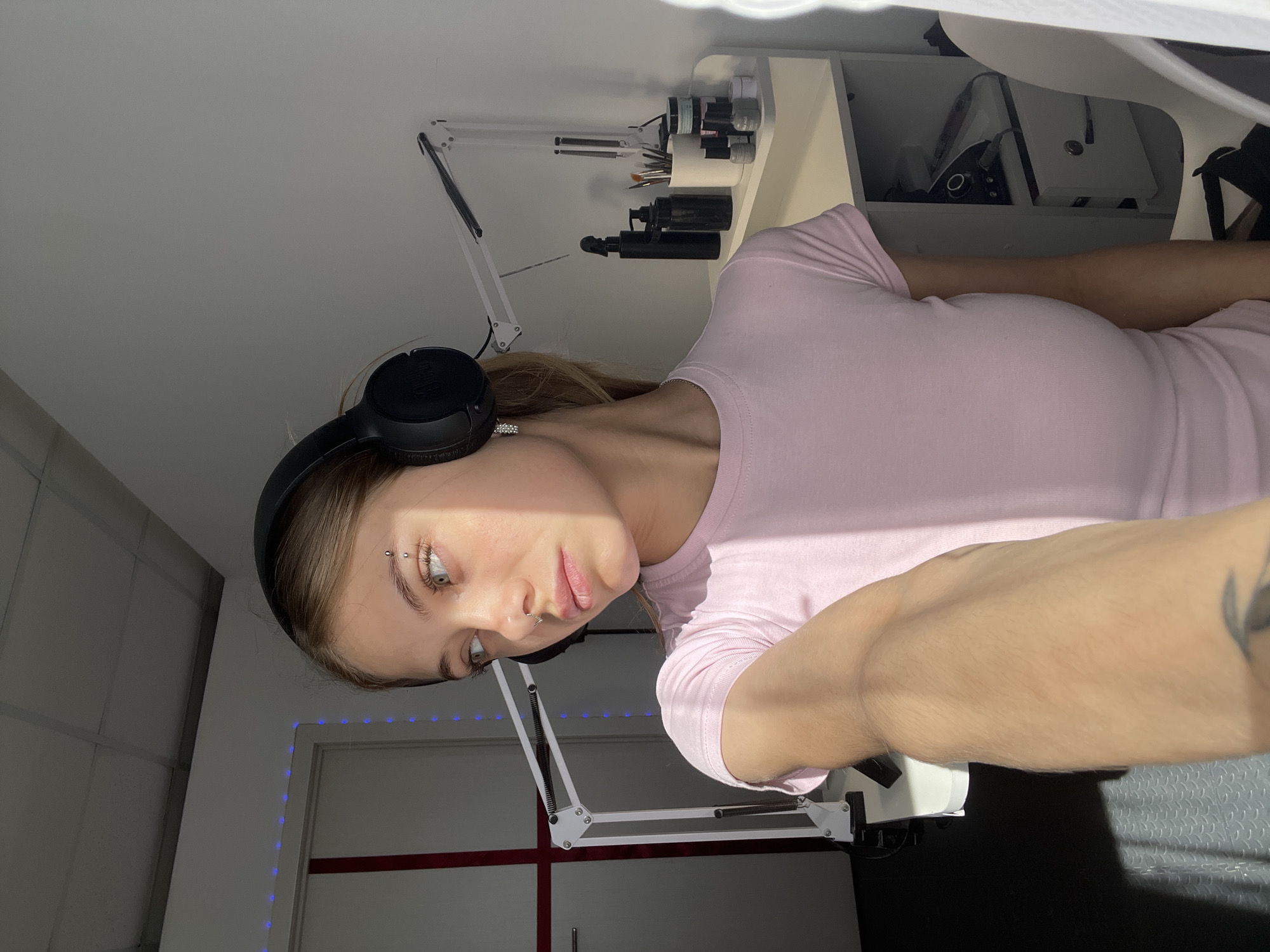A person with fair skin, light brown hair, and a piercing in their eyebrow and nose is wearing black headphones and a pink t-shirt. They are positioned near a desk with a white articulated lamp and a shelf unit holding various cosmetic products and tools. A second white articulated lamp is visible in the background, positioned against a white wall. Behind the second lamp is a white door marked with a red stripe and outlined with blue LED lights. The scene is indoors and well-lit. The exact location within Фоки, Russia is not identifiable from the image.
