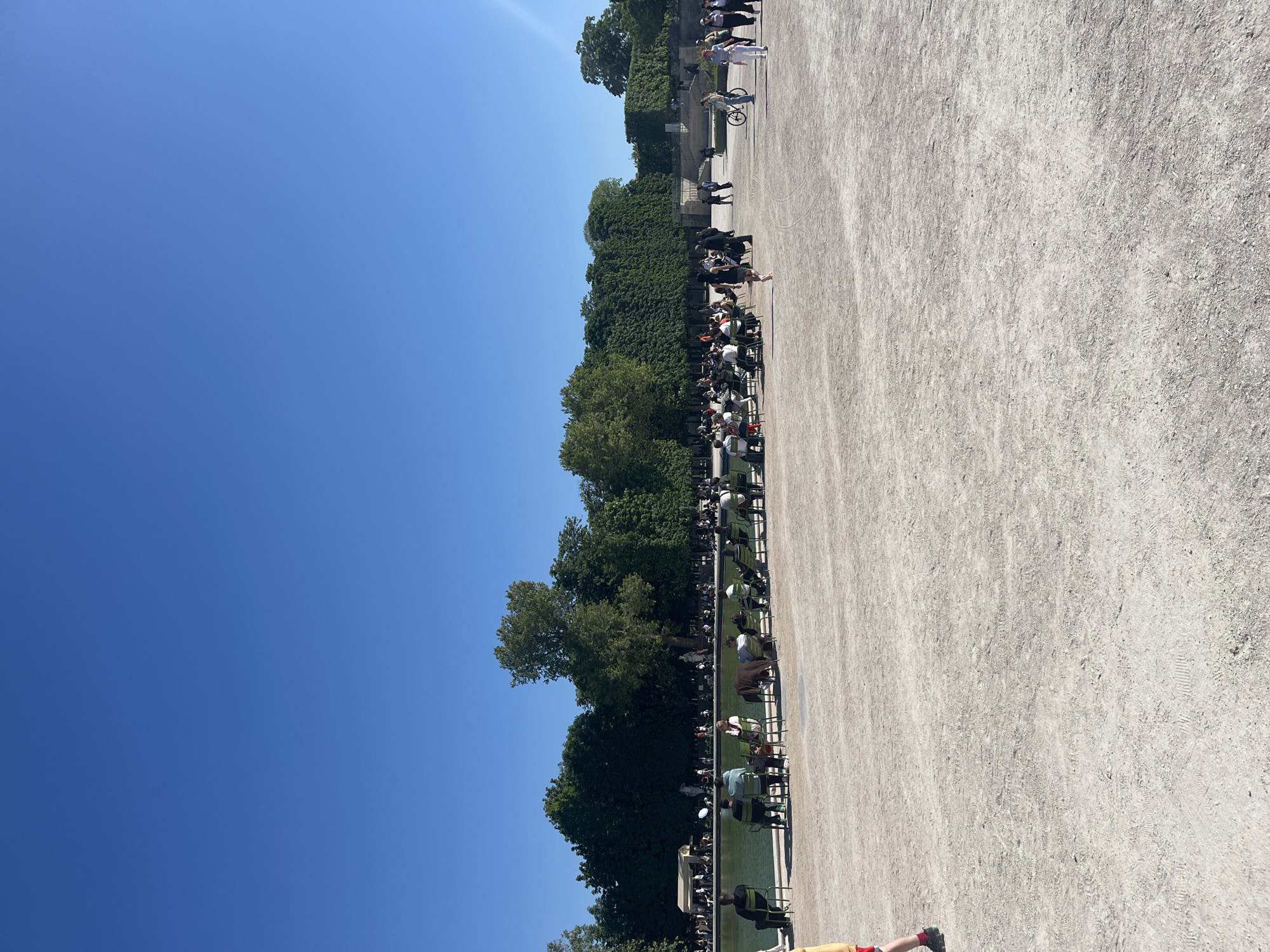 The image depicts a wide, open public park or garden under a clear, bright blue sky, typical of a sunny day in Paris, France. The scene is dominated by a light-colored gravel or dirt surface that stretches across most of the foreground and middle ground.
Along the left side of the frame, there is a long, narrow body of water, possibly a pond or canal, bordered by a large number of people. Many individuals are seated on characteristic dark green metal chairs, facing the water or towards the park. Other people are standing or strolling along the path. Beyond the water and the seated individuals, a dense line of lush green trees and manicured hedges forms a natural backdrop, suggesting the boundary of the park or a garden section. More people can be observed further back among the trees and on higher ground. A bicycle is also visible in the background.
The activity appears to be leisure and relaxation, with people enjoying the pleasant weather in a serene outdoor setting. The time of day is clearly daytime, likely midday or early afternoon, given the intensity of the sun and the distinct shadows cast on the gravel. The weather is excellent, with no clouds in the sky. The overall atmosphere conveys a sense of tranquility and enjoyment of the park. There is no visible text in the image.